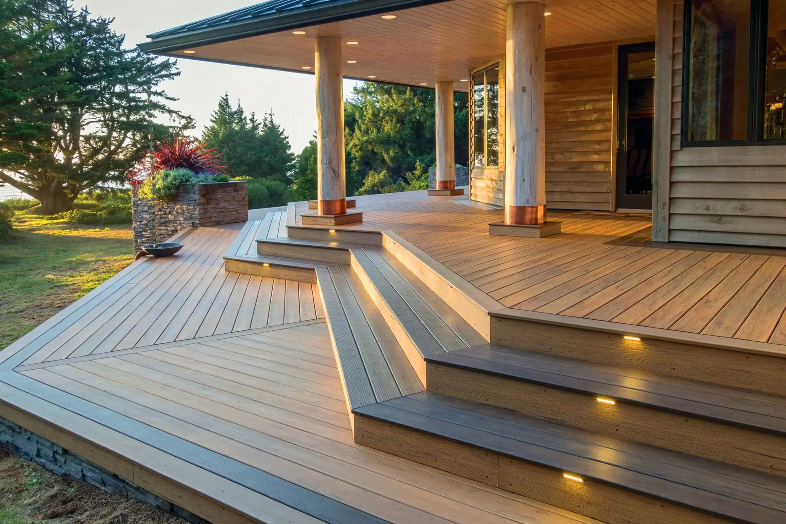 Wooden deck with steps and embedded lights, attached to a house with large windows and wooden siding, surrounded by trees and greenery during sunset.