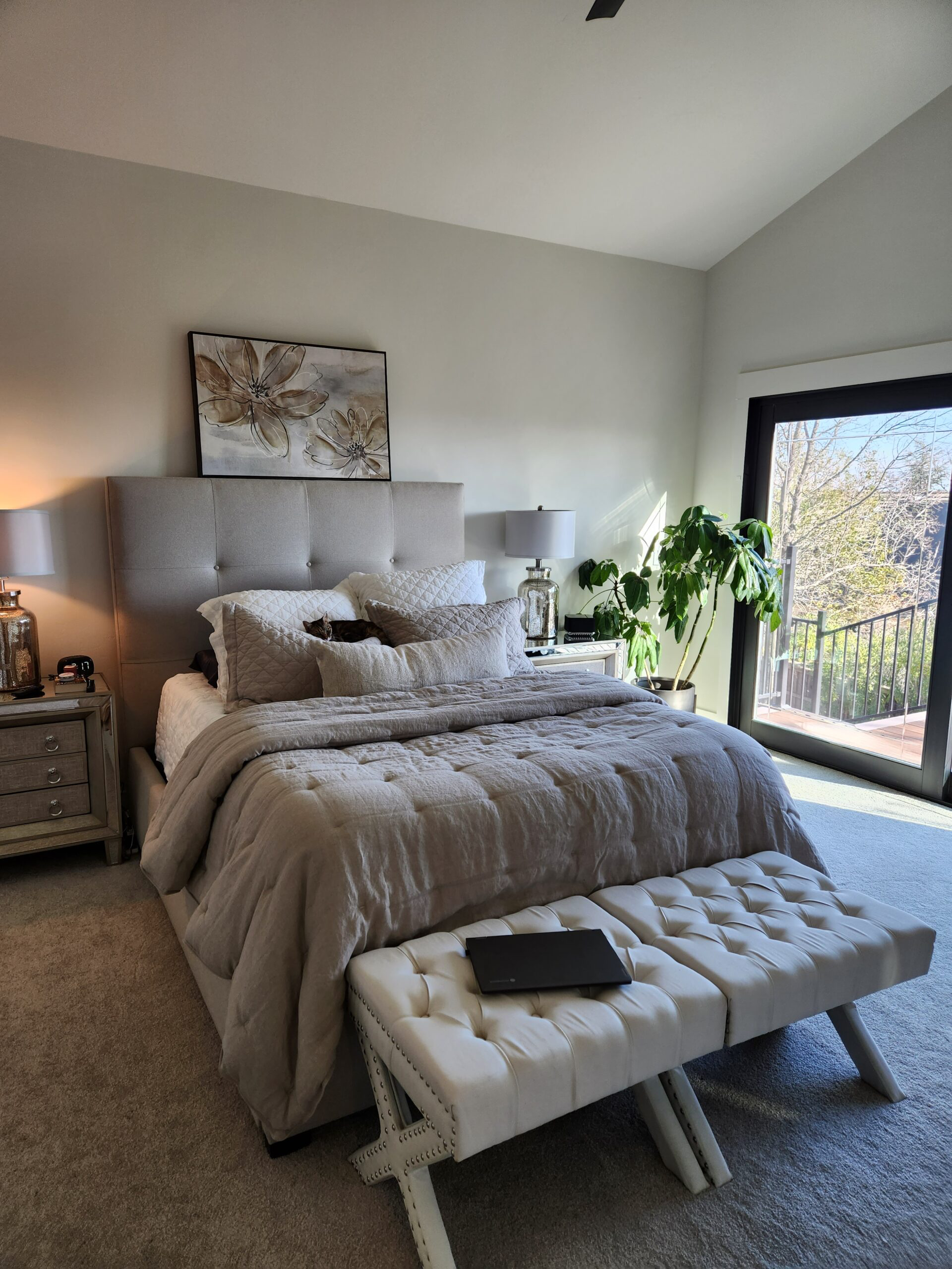 A bedroom with a bed, two nightstands with lamps, a plant near a sliding glass door, and a picture of flowers on the wall. There is a laptop on a white upholstered bench at the foot of the bed.