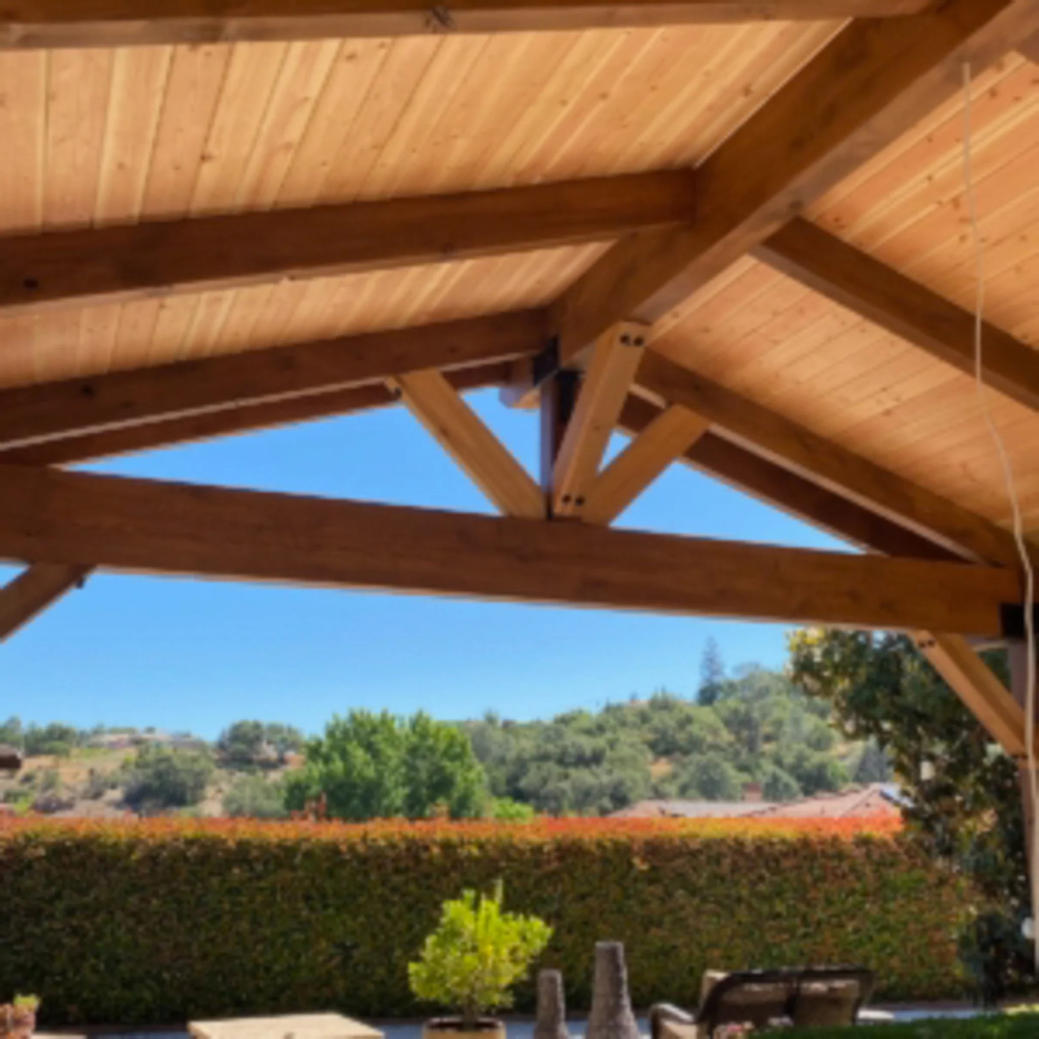 View of a wooden patio roof with exposed beams and a clear blue sky in the background, overlooking a green landscape with trees and shrubbery.