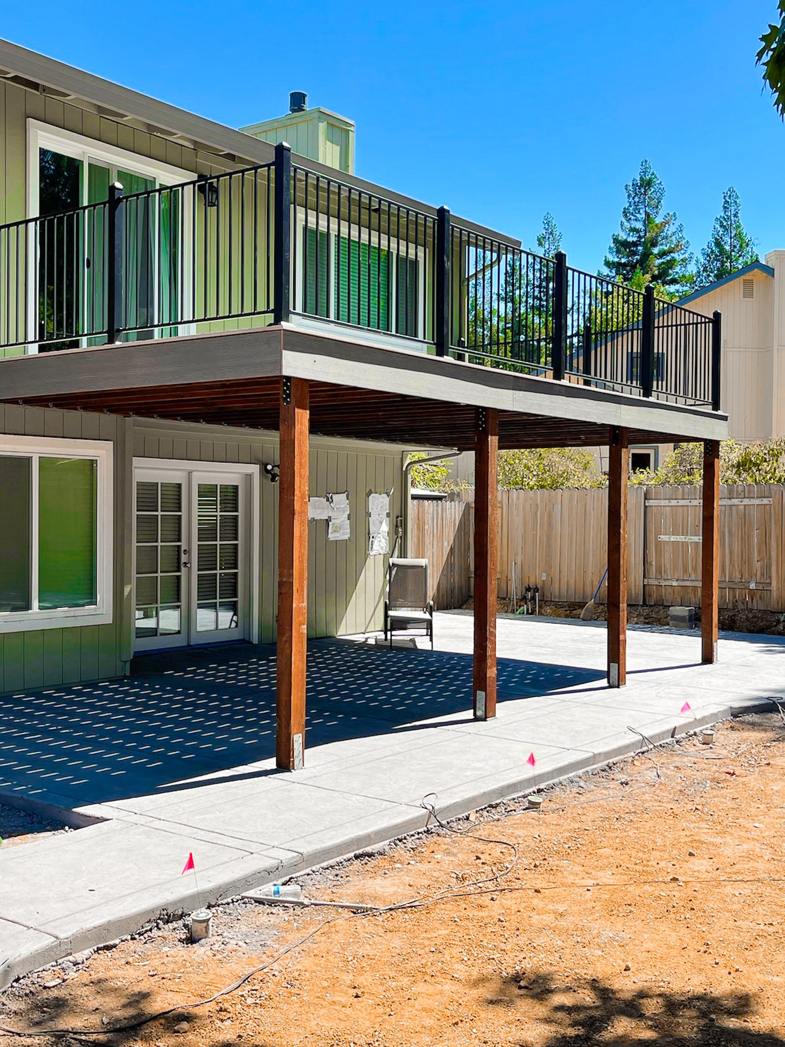 Backyard patio with concrete flooring and a raised wooden deck with black metal railing, attached to a green house with sliding glass doors and large windows, under a clear blue sky.