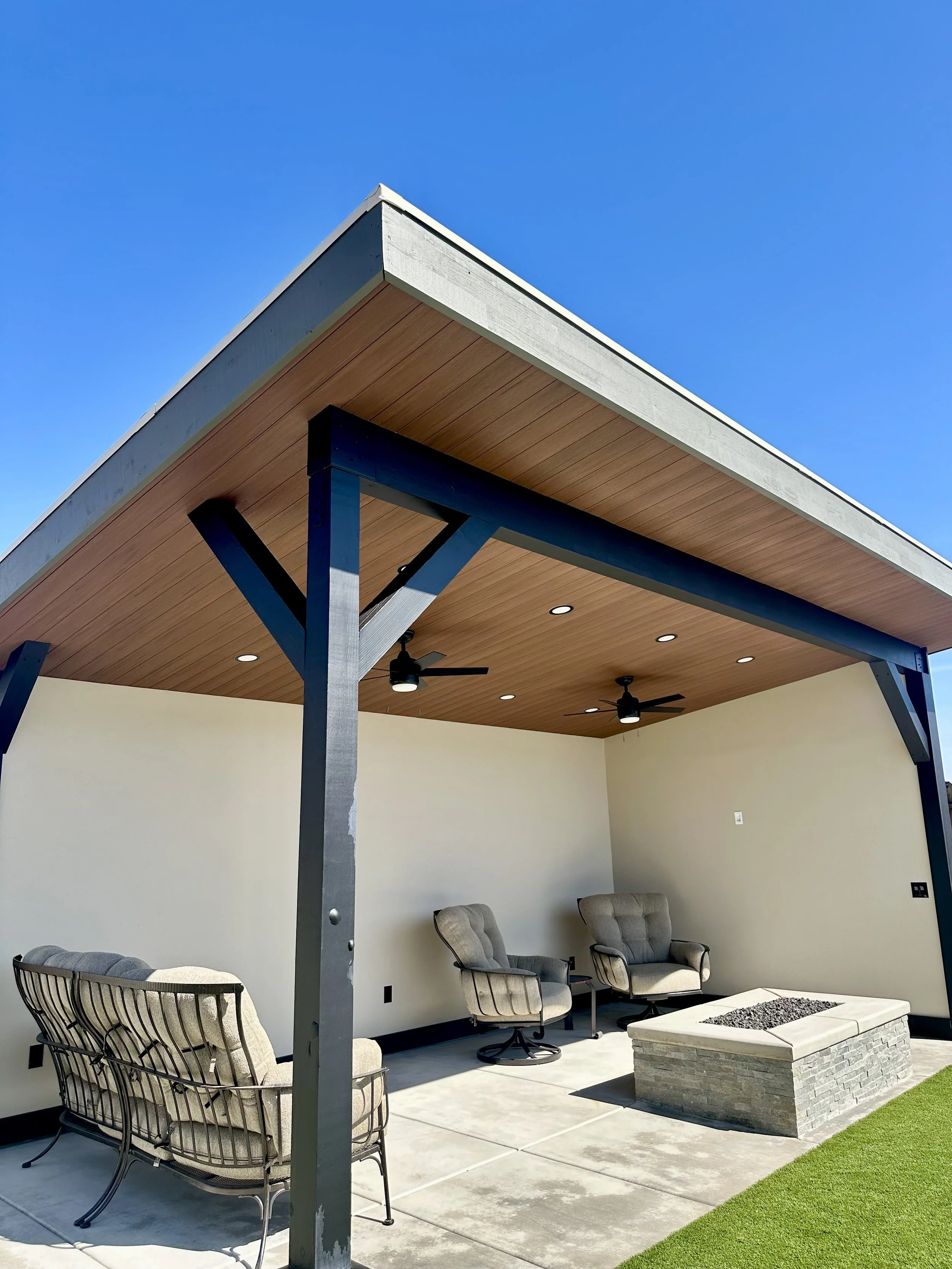 Modern outdoor patio with seating area, ceiling fans, and fire pit under a wooden ceiling with recessed lighting, set against a bright blue sky.