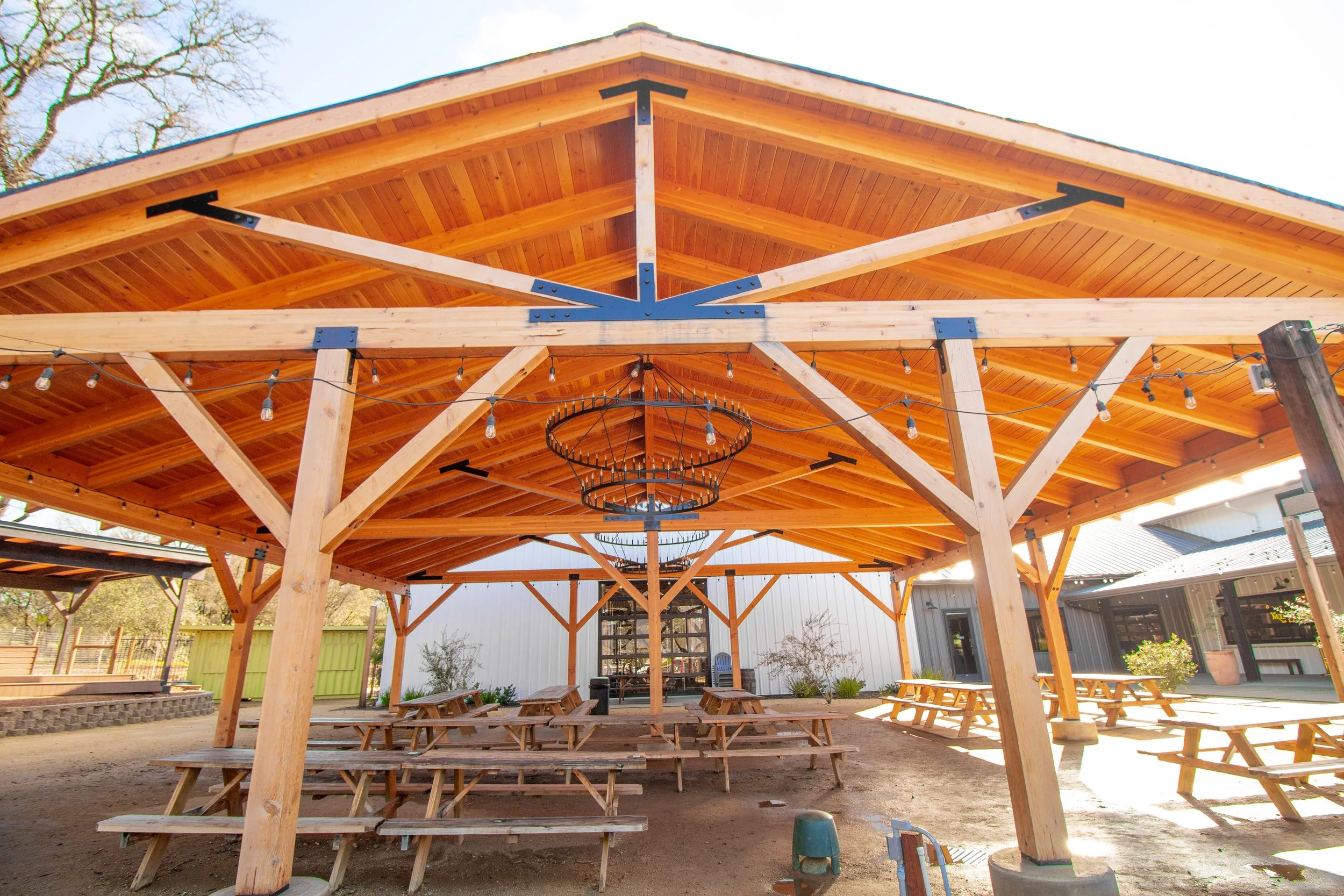 Wooden pavilion with picnic tables and string lights, outdoor setting, with trees in the background.