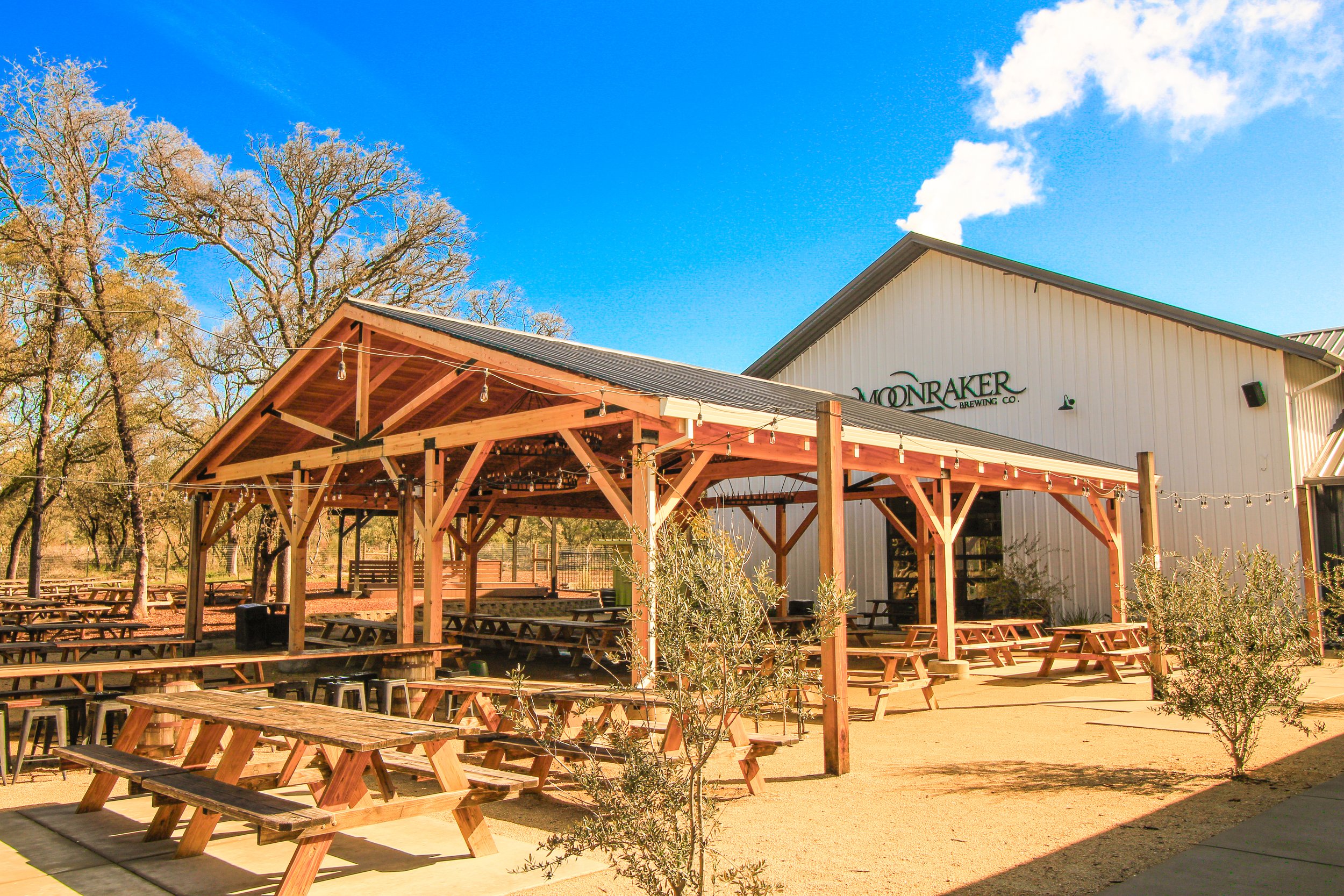 Outdoor patio with wooden picnic tables and a covered seating area at Monraker Brewing Co., with a clear blue sky and leafless trees in the background.
