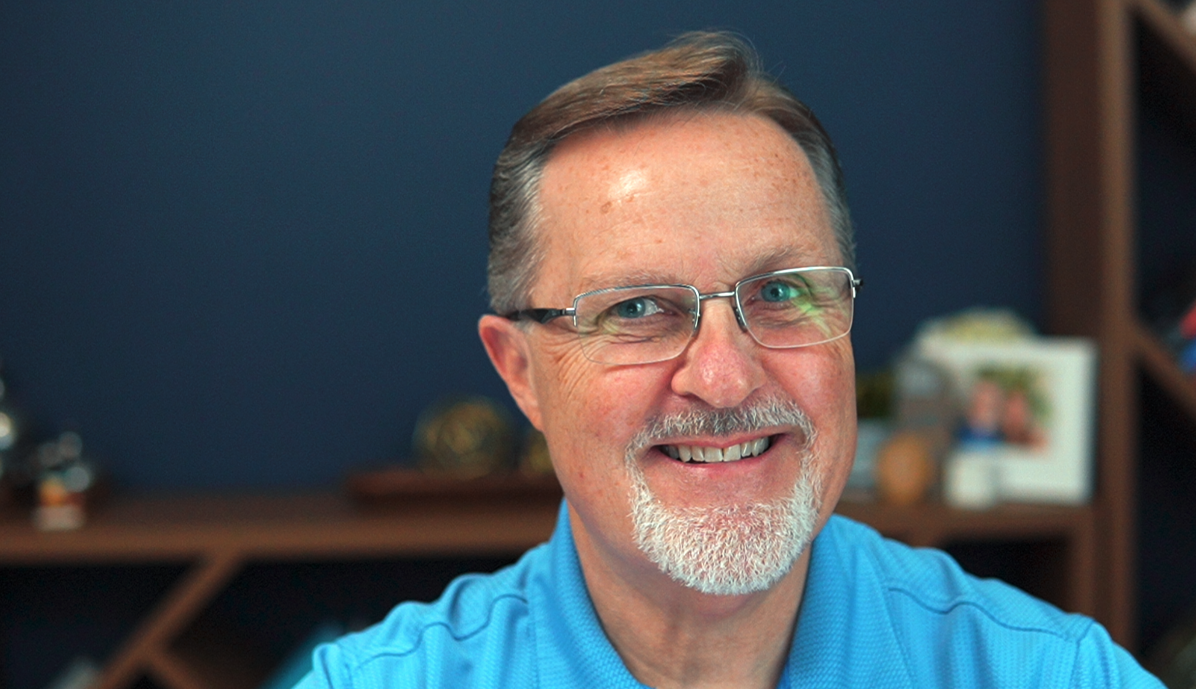 A middle-aged man with glasses, a beard, and a smile is wearing a blue polo shirt, sitting in a room with a bookshelf and personal items in the background.