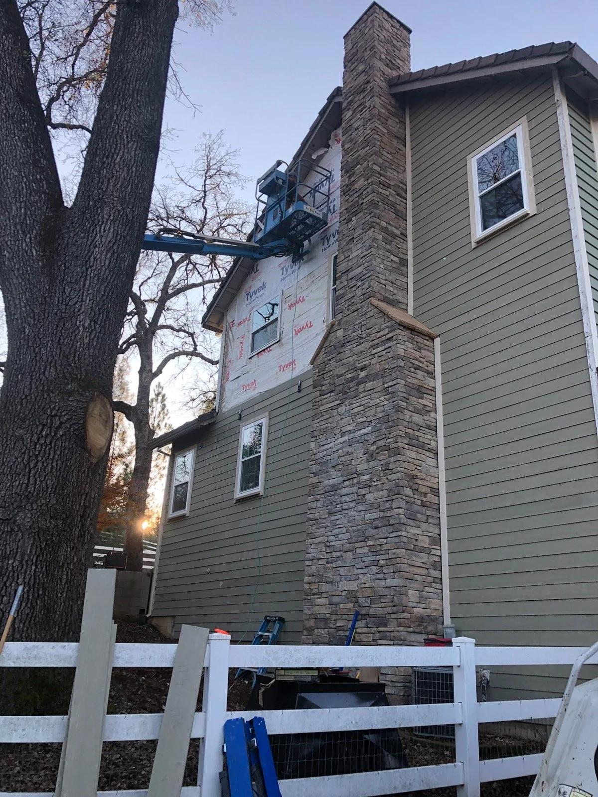 A house undergoing exterior renovations with a chimney and partial siding. A construction lift is positioned near the house, and scaffolding or equipment are visible near the ground. The house has a stone chimney and new siding on part of the exterio
