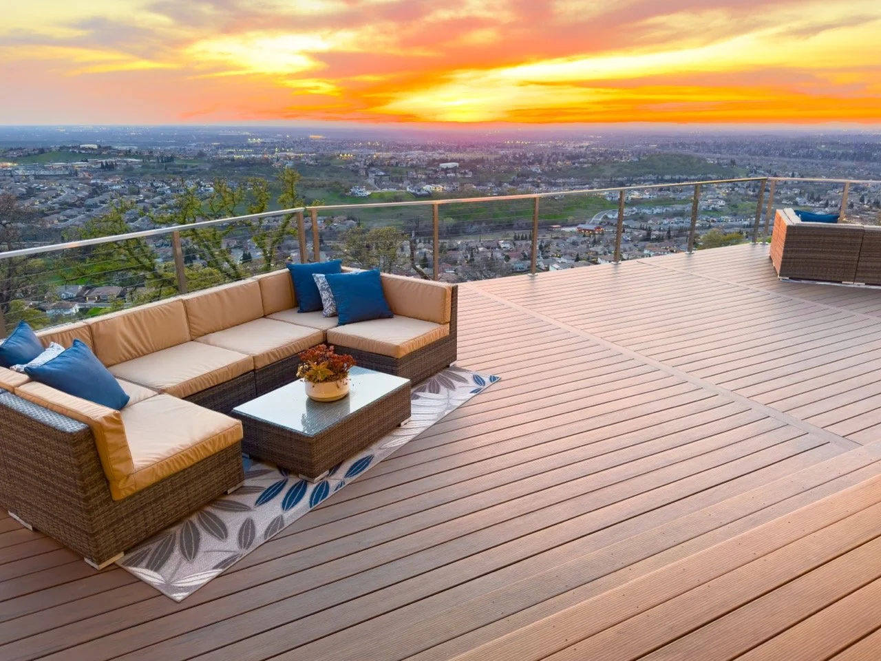 Outdoor rooftop patio with sectional sofa, blue cushions, and coffee table overlooking a cityscape at sunset.