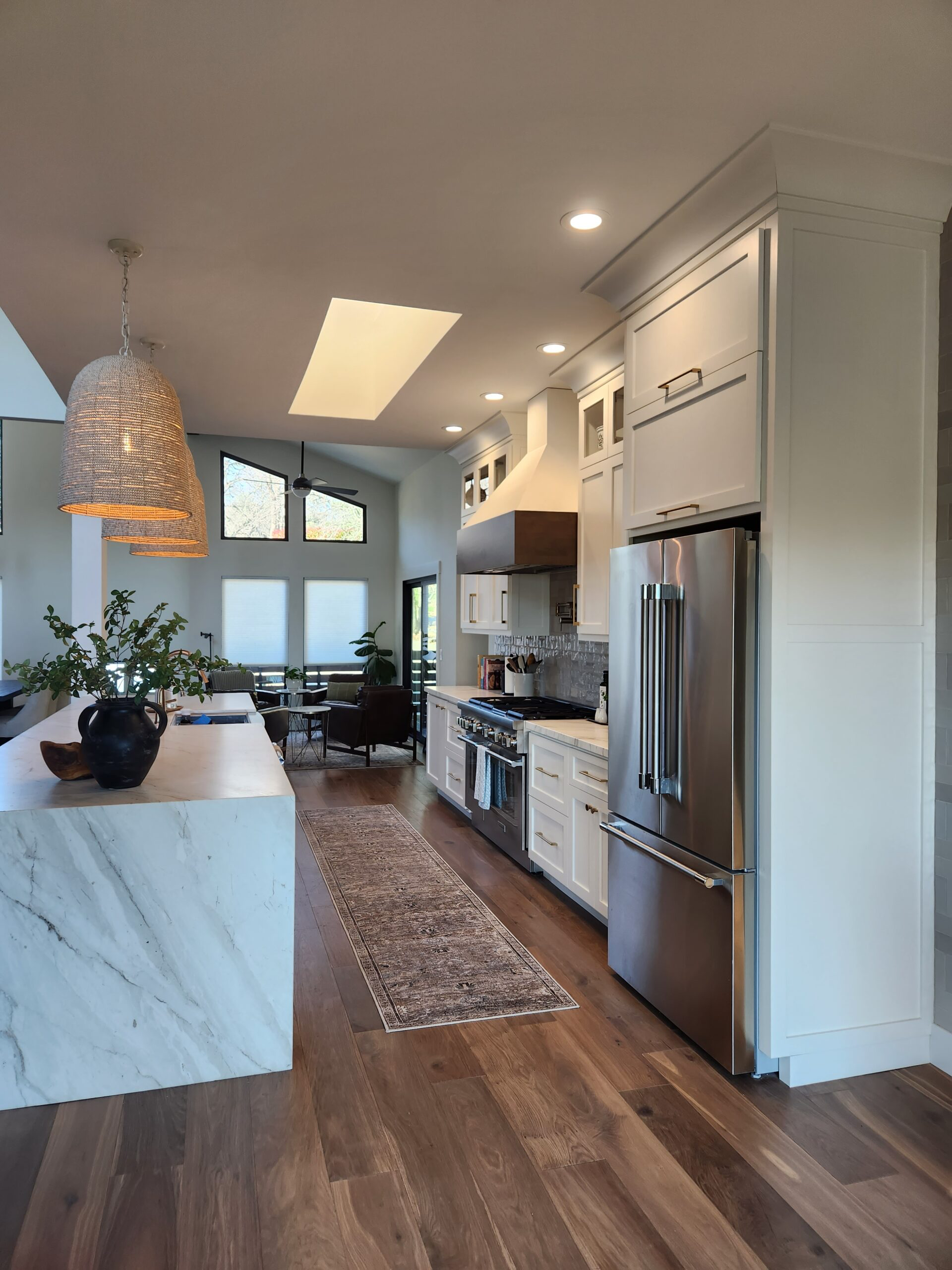 Modern kitchen with white cabinets, stainless steel refrigerator, and a marble island. In the background, there is a living area with large windows and seating.