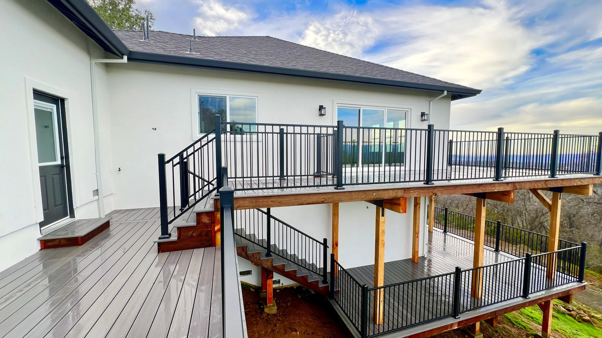 Newly built multi-level outdoor deck with black metal railings and wooden support beams, attached to a white house with sliding glass doors and a nearby black door.