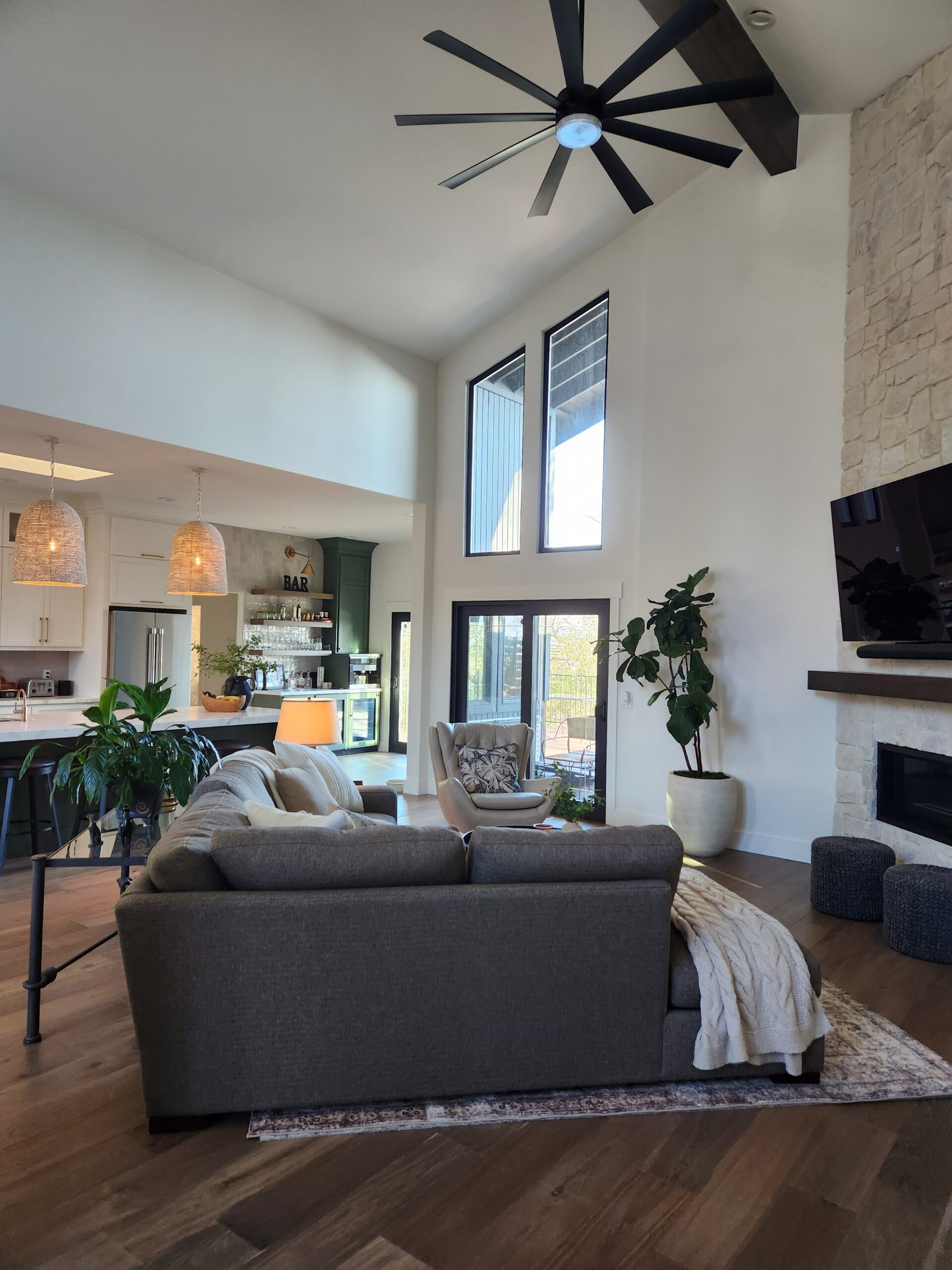 Living room with high ceiling, large windows, ceiling fan, grey sofa, armchair, fireplace, TV, potted plant, and sliding glass door leading to porch.