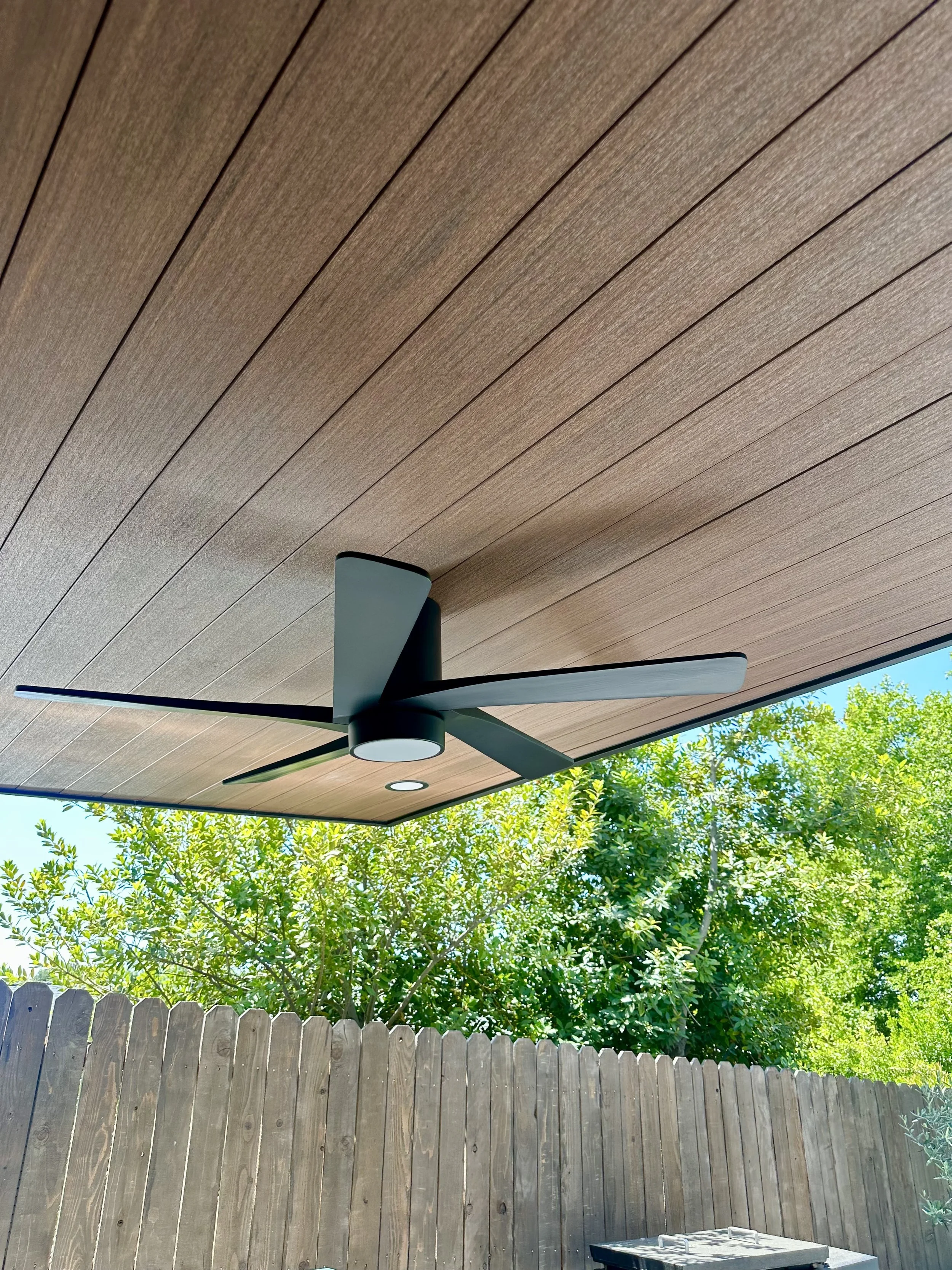 Outdoor patio with wooden ceiling, ceiling fan, and a wooden fence with green trees in the background.
