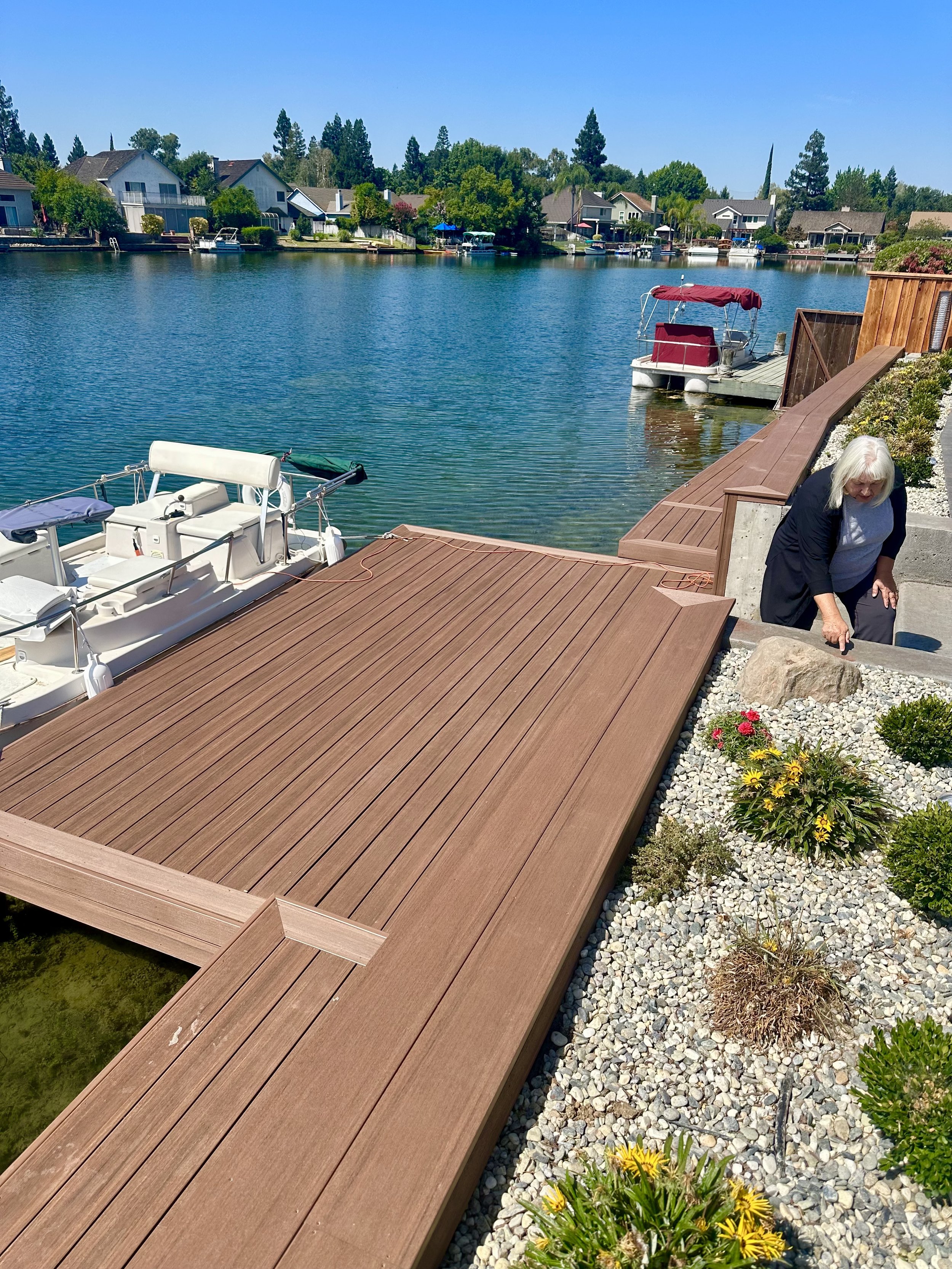 Woman gardening beside a houseboat dock on a calm lake, with houses and boats visible across the water under a clear blue sky.