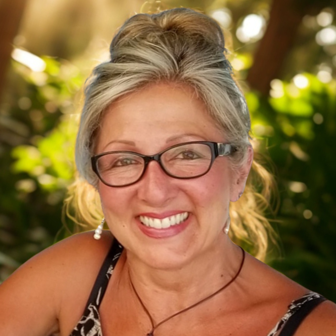 A woman with glasses and earrings smiling outdoors with greenery and sunlight in the background.