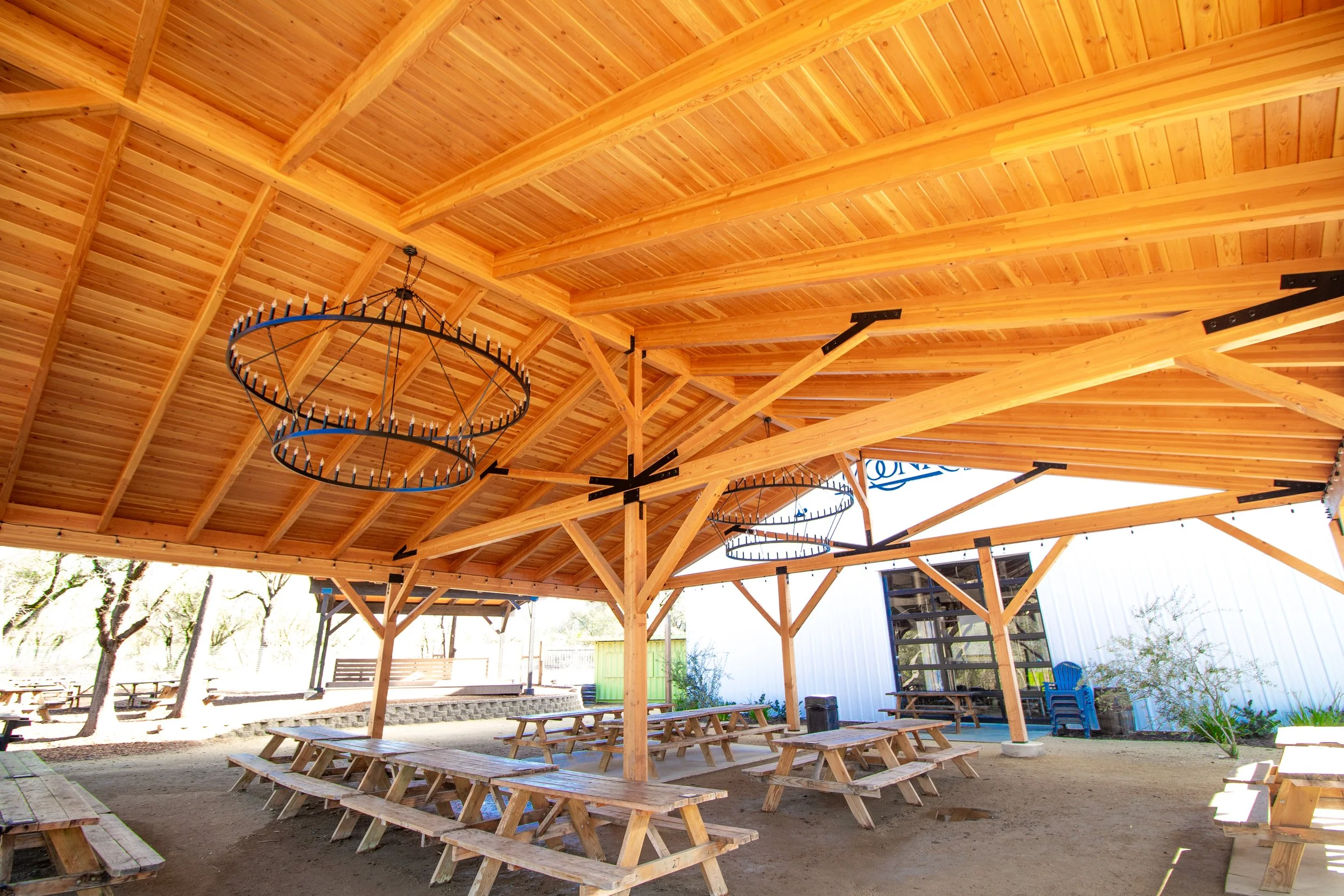 Open-air pavilion with wooden roof and tables, decorative lighting fixtures hanging from the ceiling, surrounding trees, and outdoor seating.