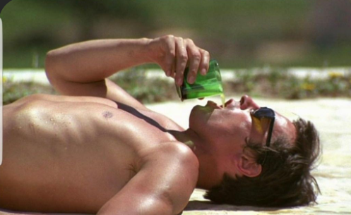 A man lying on the ground outdoors, drinking from a green can.