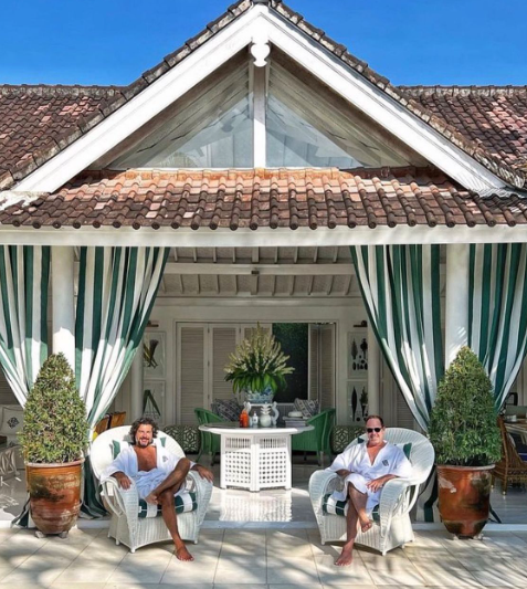 Two men relaxing on patio chairs in front of a white house with large green and white curtains and potted plants.
