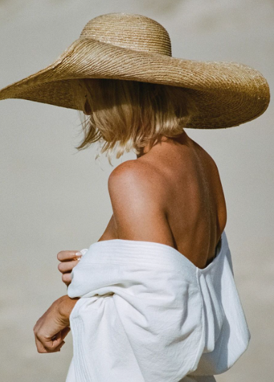 Person on the beach wearing a large straw hat and a white towel or cover-up, with their back to the camera.