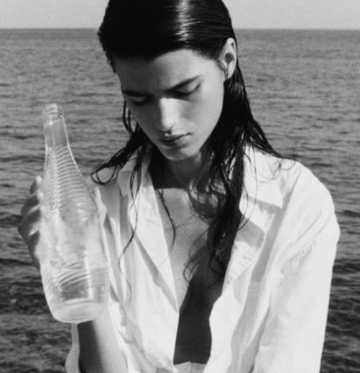 A young woman with wet hair looking down while holding an empty plastic bottle at the beach.
