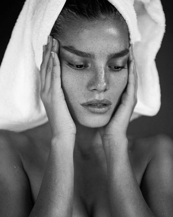 A woman with a towel wrapped around her head, touching her face with her hands, in black and white.