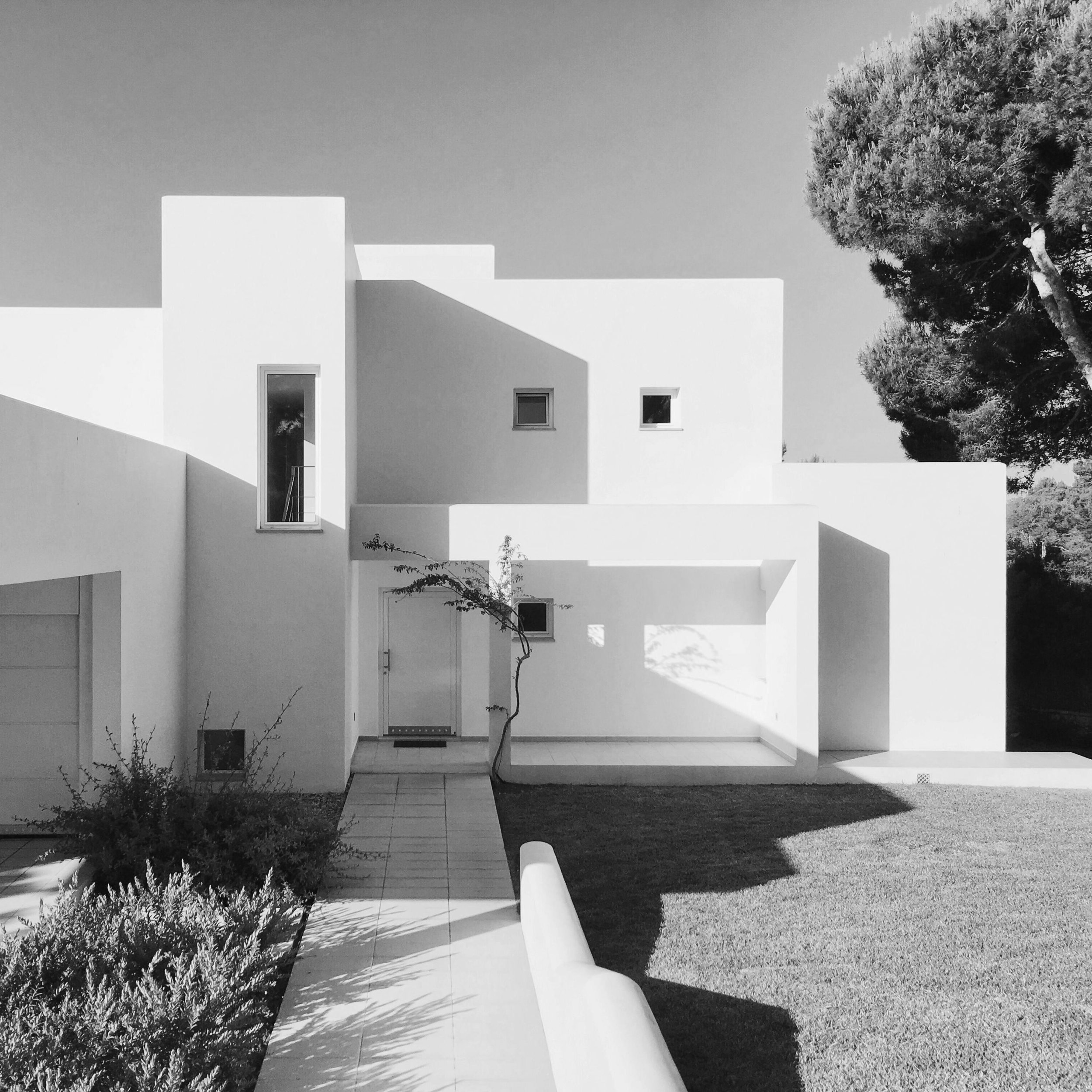 Modern white building with geometric architectural design, small landscape trees, and a manicured lawn in sunlight.