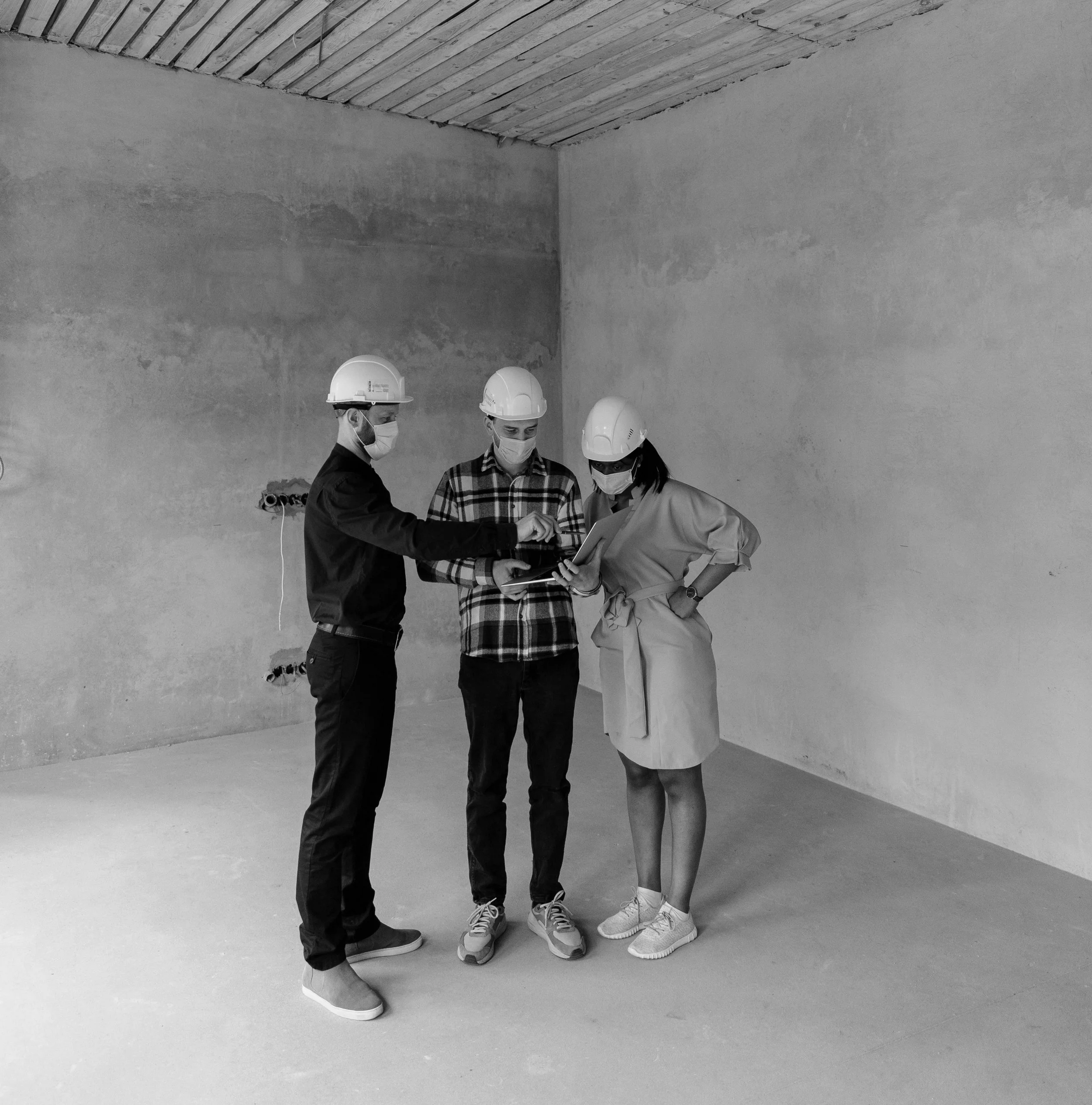 Three construction workers, wearing hard hats and masks, reviewing plans or documents inside an unfinished building.