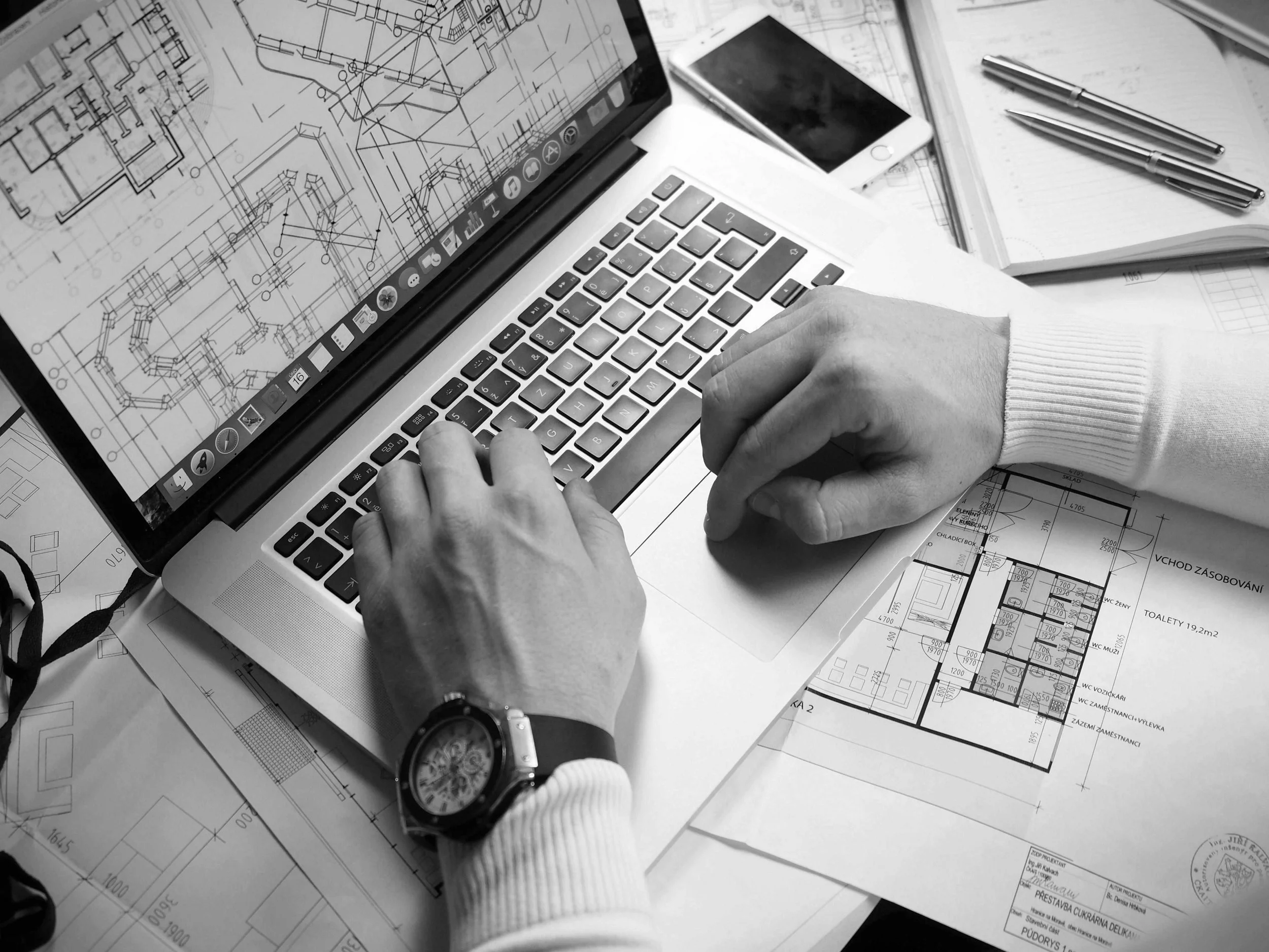 A person working on architectural plans on a laptop surrounded by blueprints, a smartphone, notebooks, and pens.