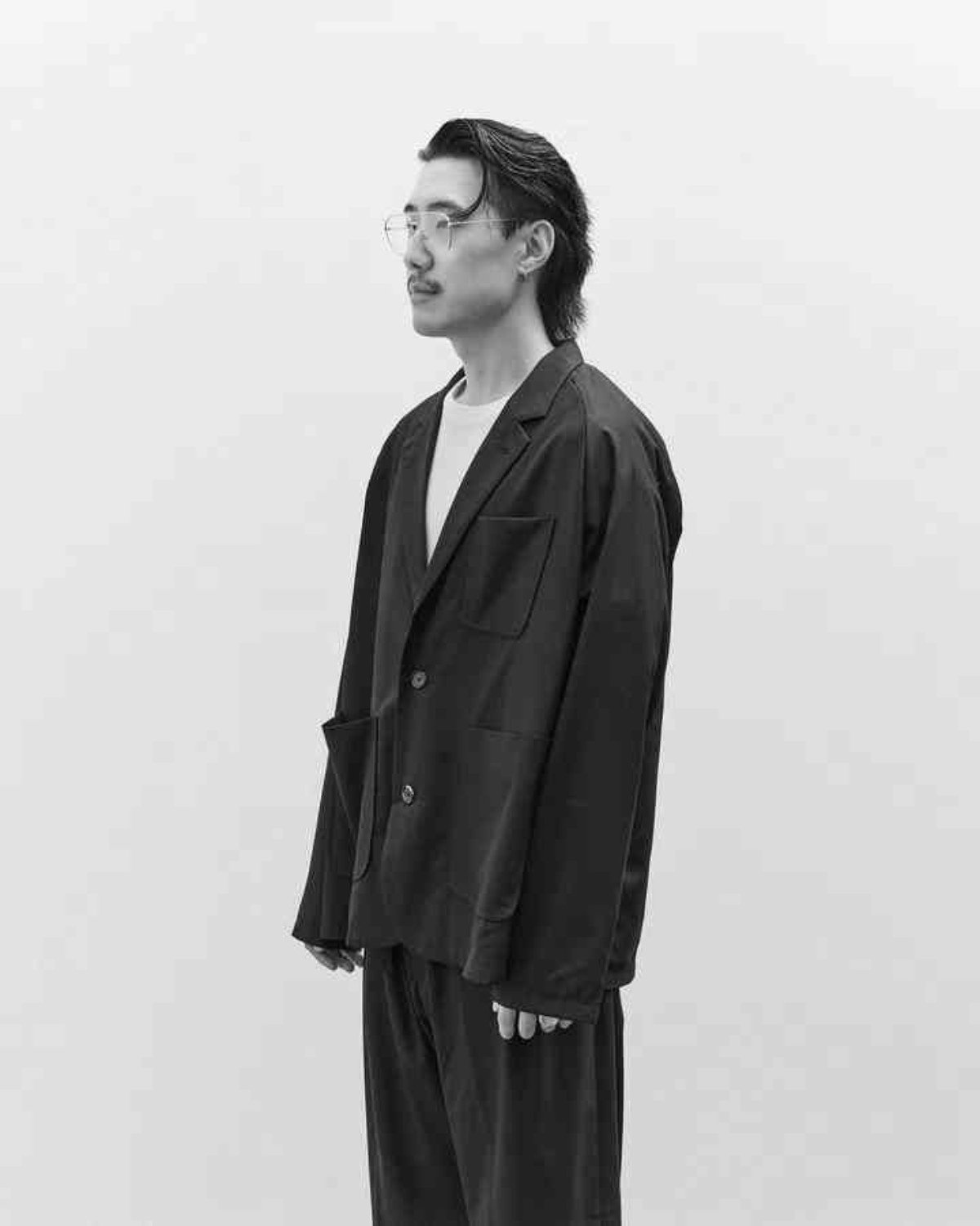 A man with glasses and slicked-back hair wearing a dark blazer and pants, standing in profile against a plain background.