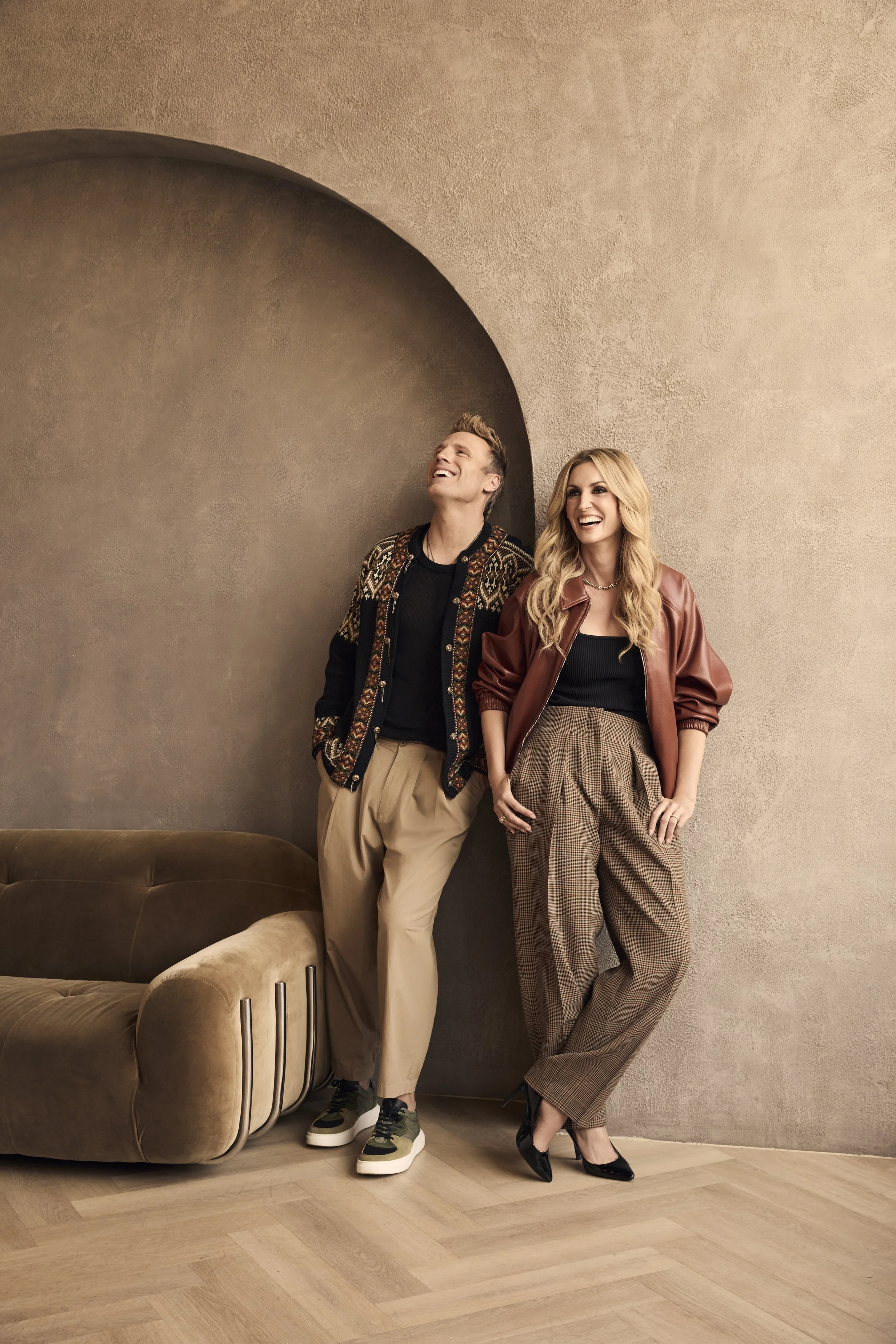 A man and woman standing and leaning against a textured beige wall, smiling and laughing, with a brown sofa nearby.