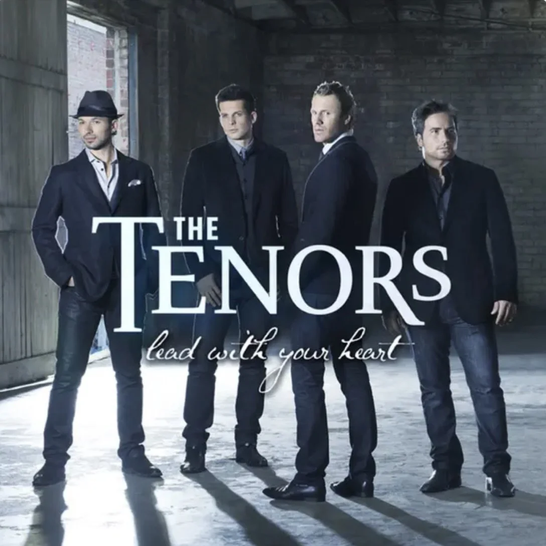 A man in a fedora hat and suit stands along with four other men in suits inside a dimly lit room with brick walls. The text on the image reads 'The Tenors' with a tagline 'lead with your heart'.