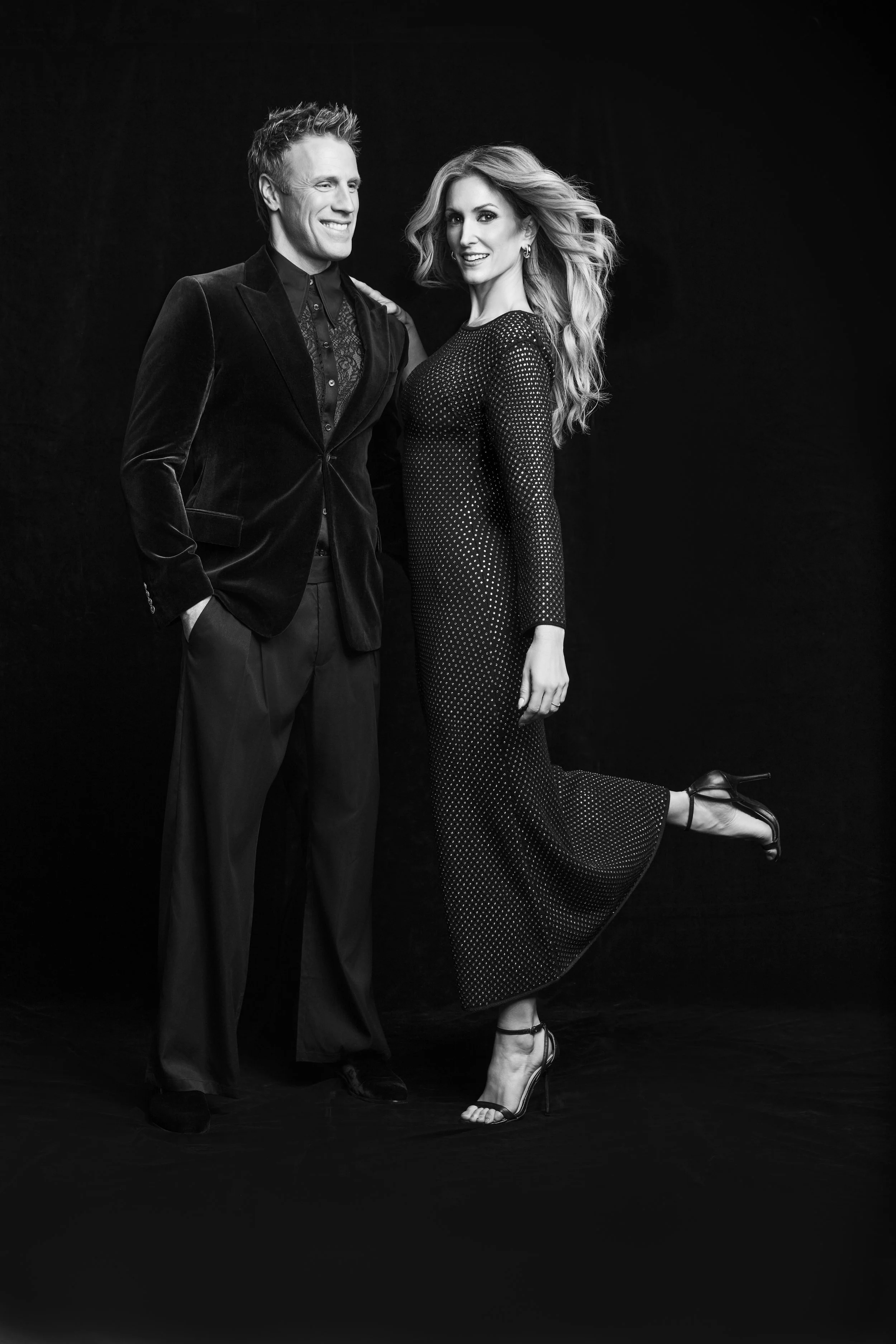 Black and white photo of a man and a woman standing together against a dark background, dressed in formal attire, smiling and posing.