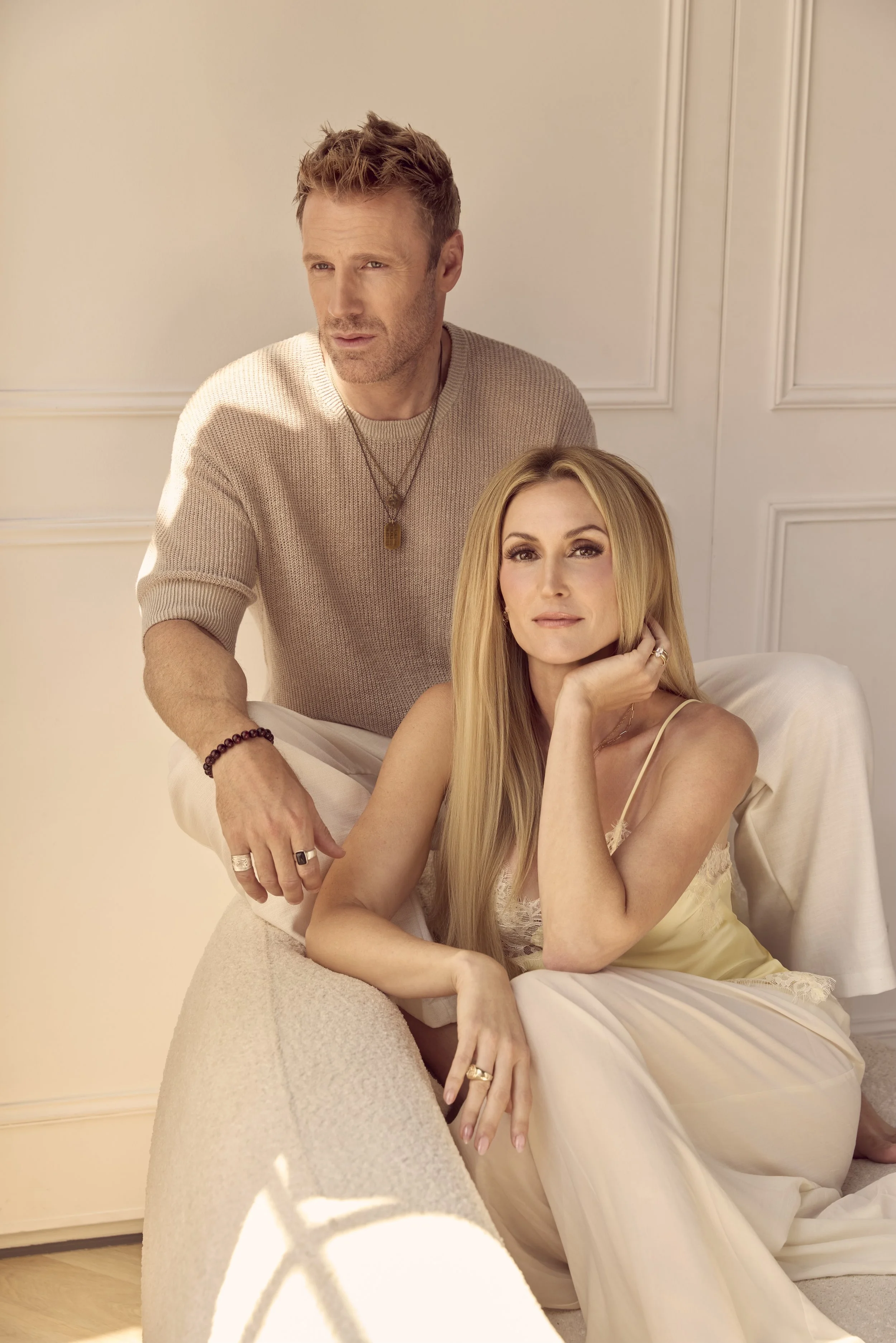 A man and a woman posing indoors. The man is standing behind the woman, who is sitting on a couch. The woman has long blonde hair and is wearing a light-colored slip dress. The man has short, tousled hair, wearing a beige sweater, and multiple rings 