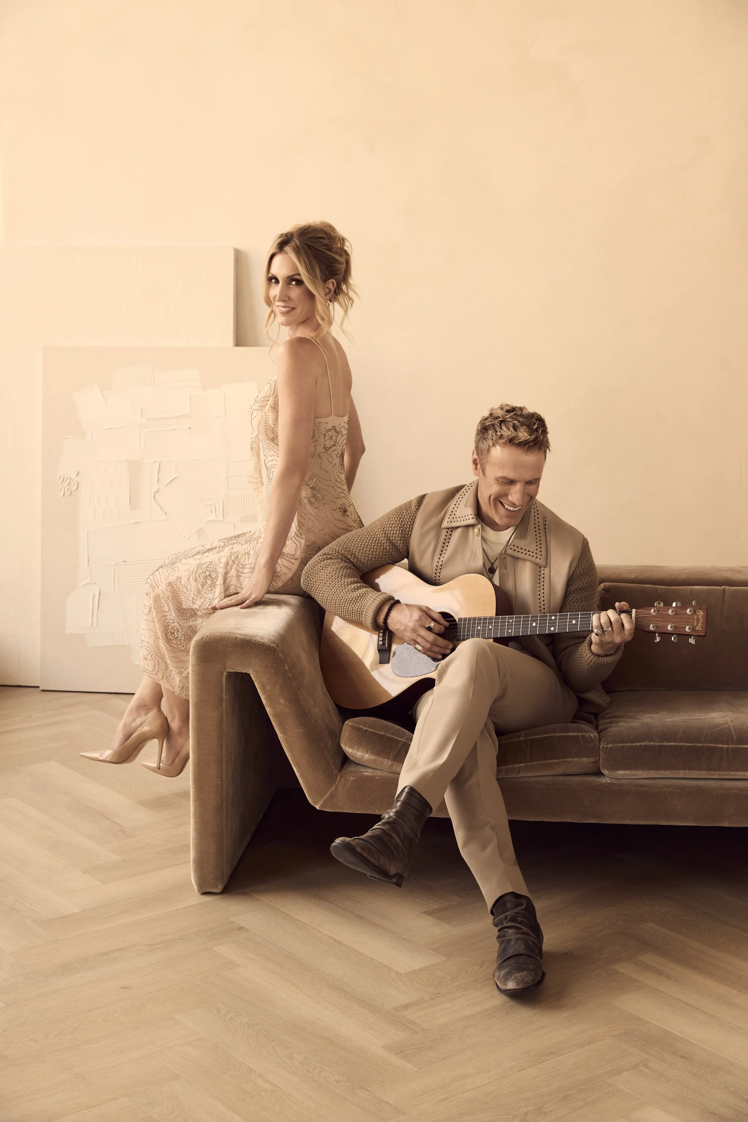 A woman in a lace dress and heels sitting on the back of a brown couch, smiling, while a man in beige pants and boots plays an acoustic guitar and sits on the couch, both appearing happy in a cozy, softly lit room.