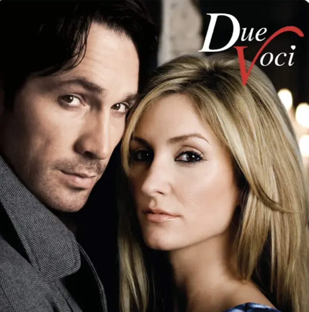 A close-up of a man and a woman with serious expressions, standing closely together. The man has dark hair and the woman has long blonde hair. There is text in the upper right corner that reads 'Due Voci'.