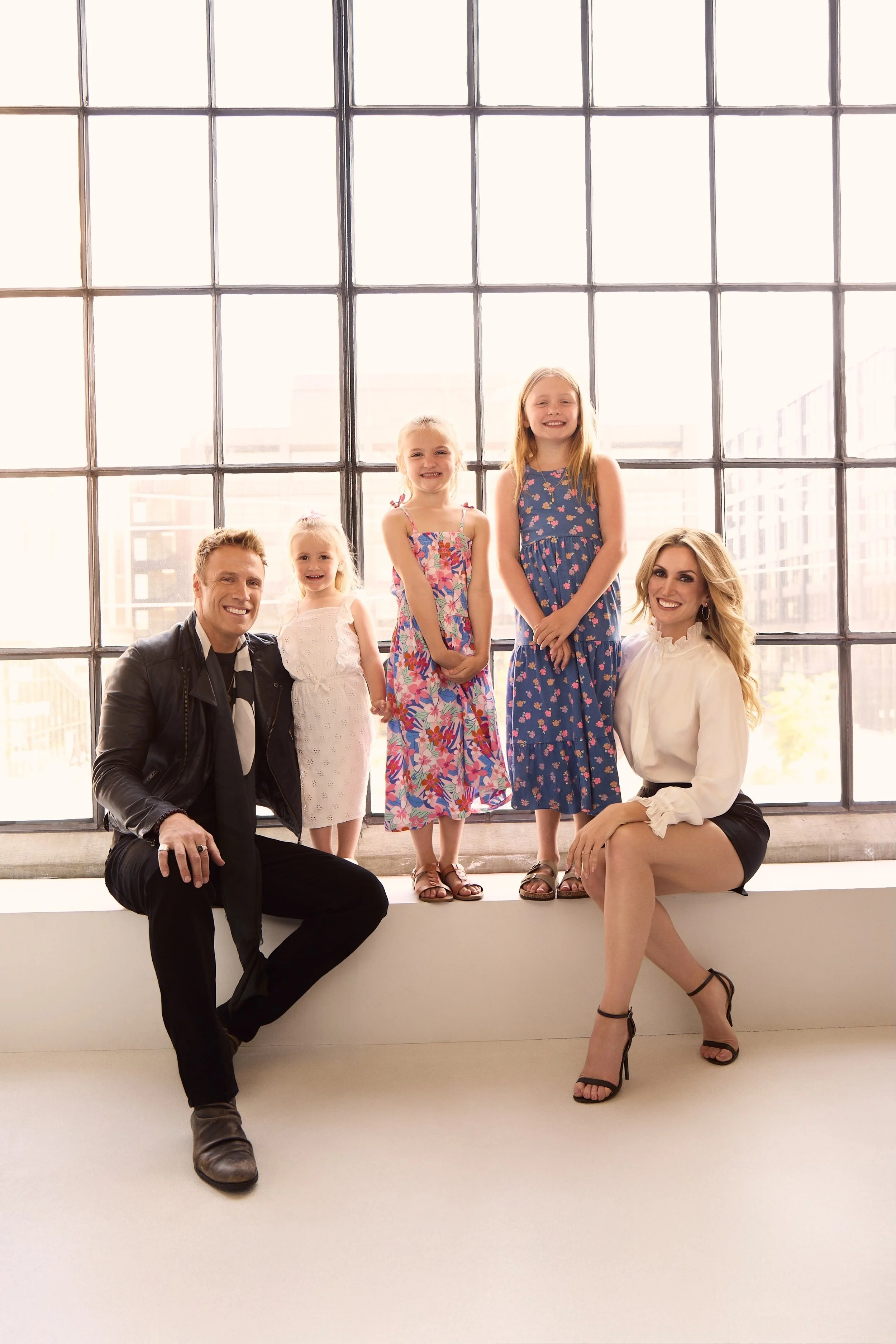 A family posing in front of a large window with city buildings outside. Two adults, three young girls The man is wearing a leather jacket, the woman a white blouse and black shorts. The children are in colorful dresses and one is in all white