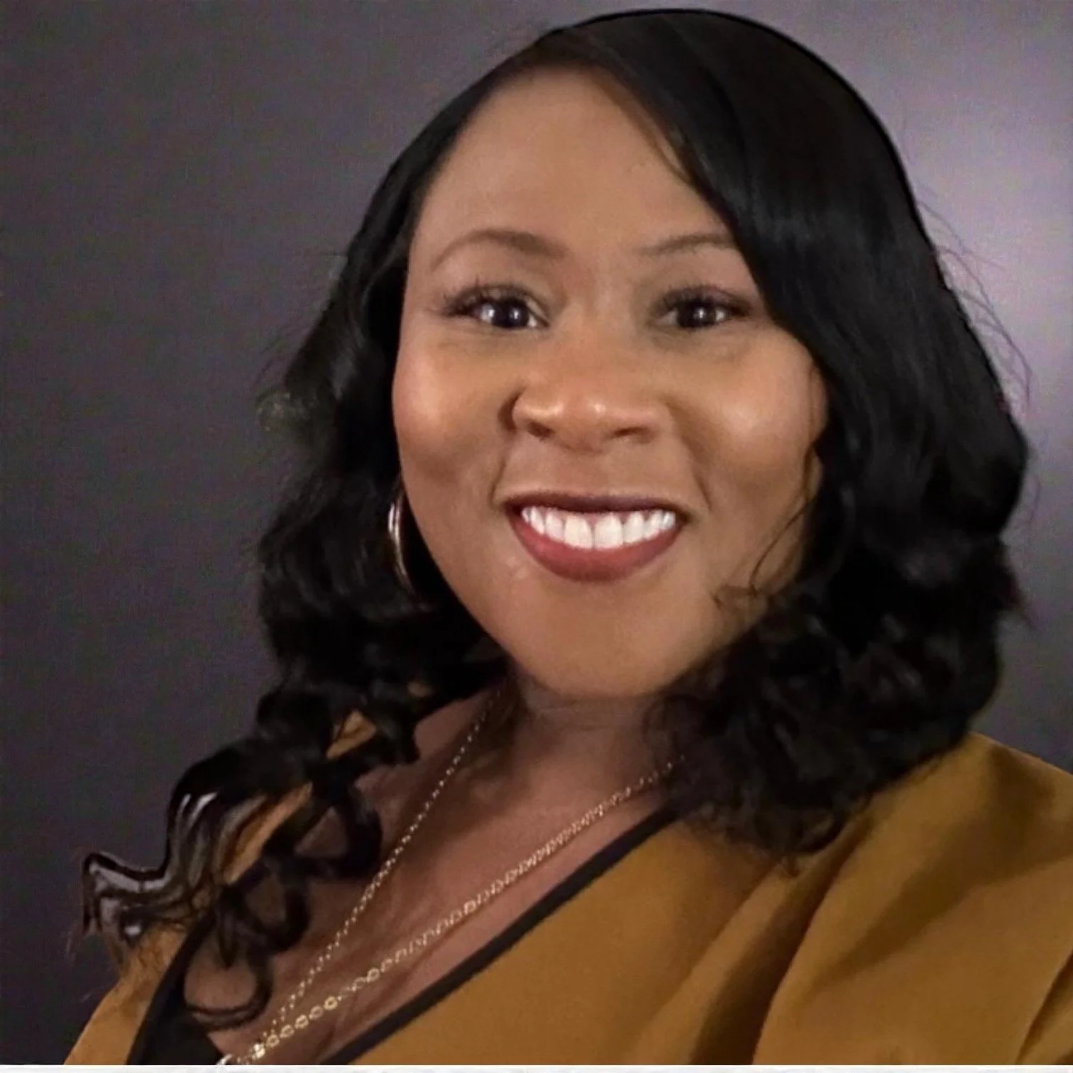 A woman with dark, curly hair styled in loose waves, smiling with white teeth, wearing a mustard yellow top, gold hoop earrings, and layered necklaces, against a dark background.