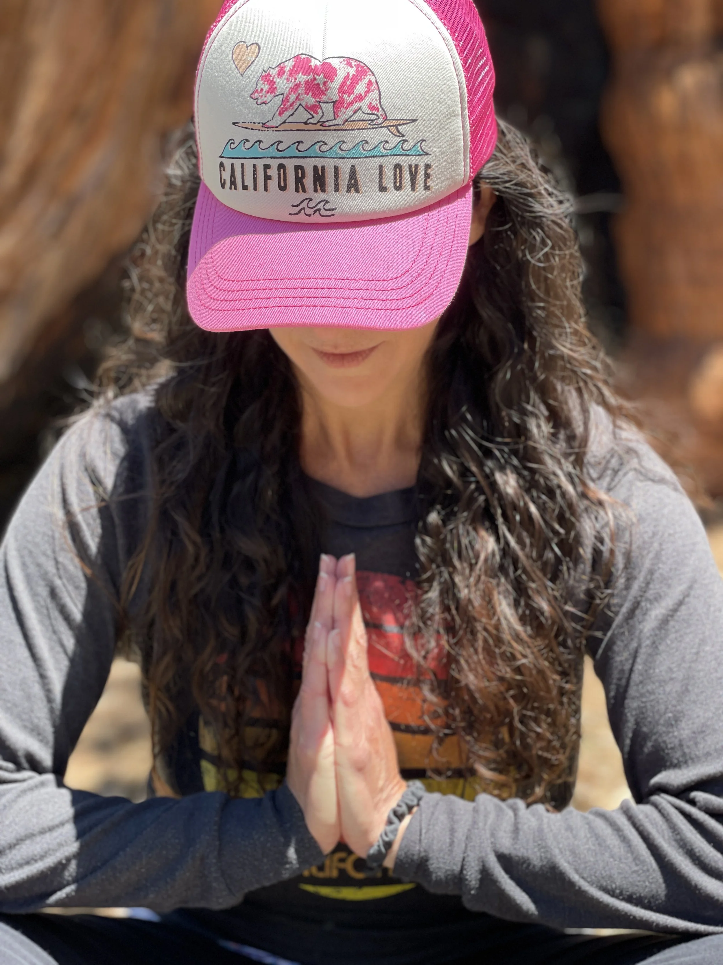 A person with long curly brown hair and a gray long sleeve shirt is doing yoga or meditation outdoors, with their hands pressed together in prayer position, and wearing a pink and white trucker hat with a bear illustration and the words "California Love" on it.