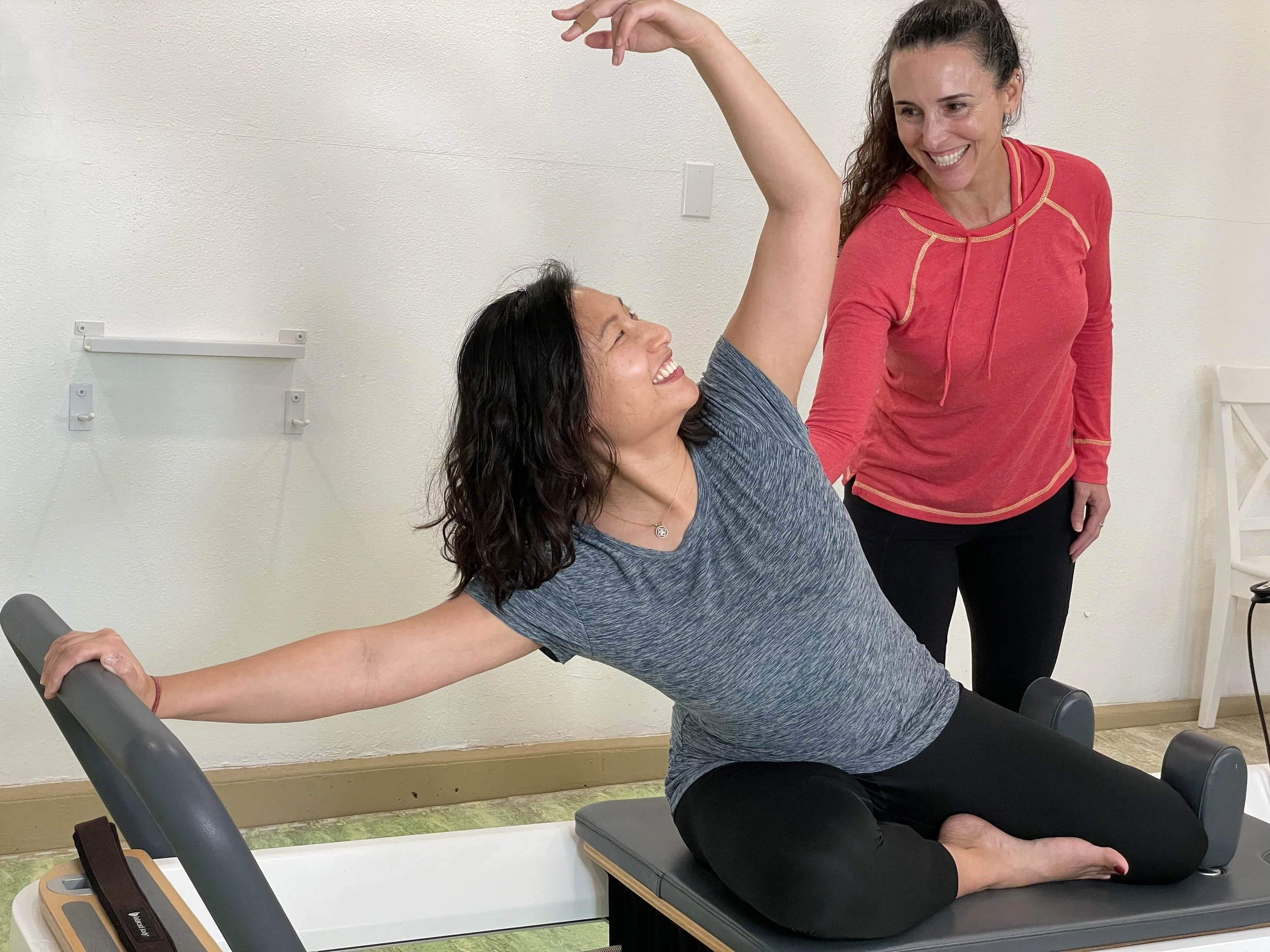 Two women are practicing Pilates in a studio. One woman, with dark wavy hair and a blue t-shirt, is in a seated stretch on a Pilates reformer machine, smiling and looking at the other woman. The other woman, with curly dark hair and a red hoodie, is standing beside her, smiling and looking at her yoga partner.