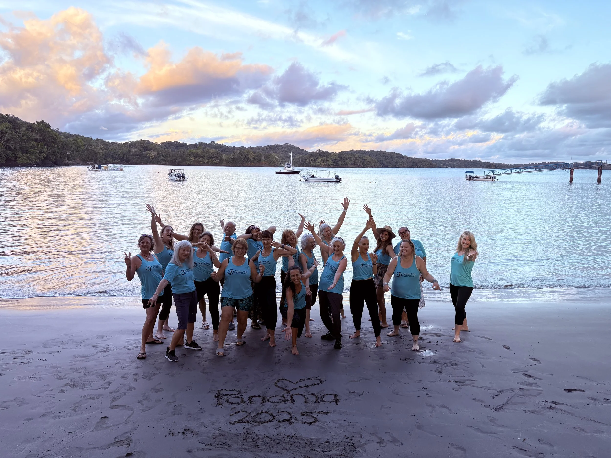 A group of smiling people wearing blue shirts at the beach during sunset, with boats on the water and a cloudy sky in the background.