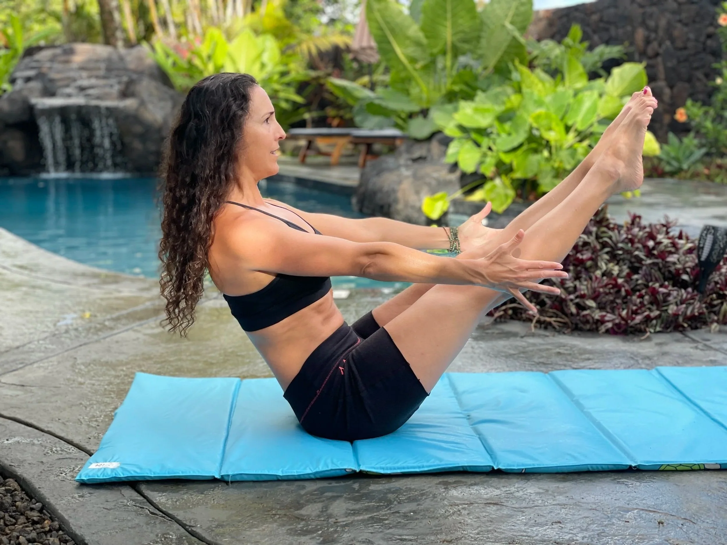 A woman with long curly hair in a black sports bra and shorts practicing yoga on a blue mat outdoors, surrounded by lush tropical plants and rocks.