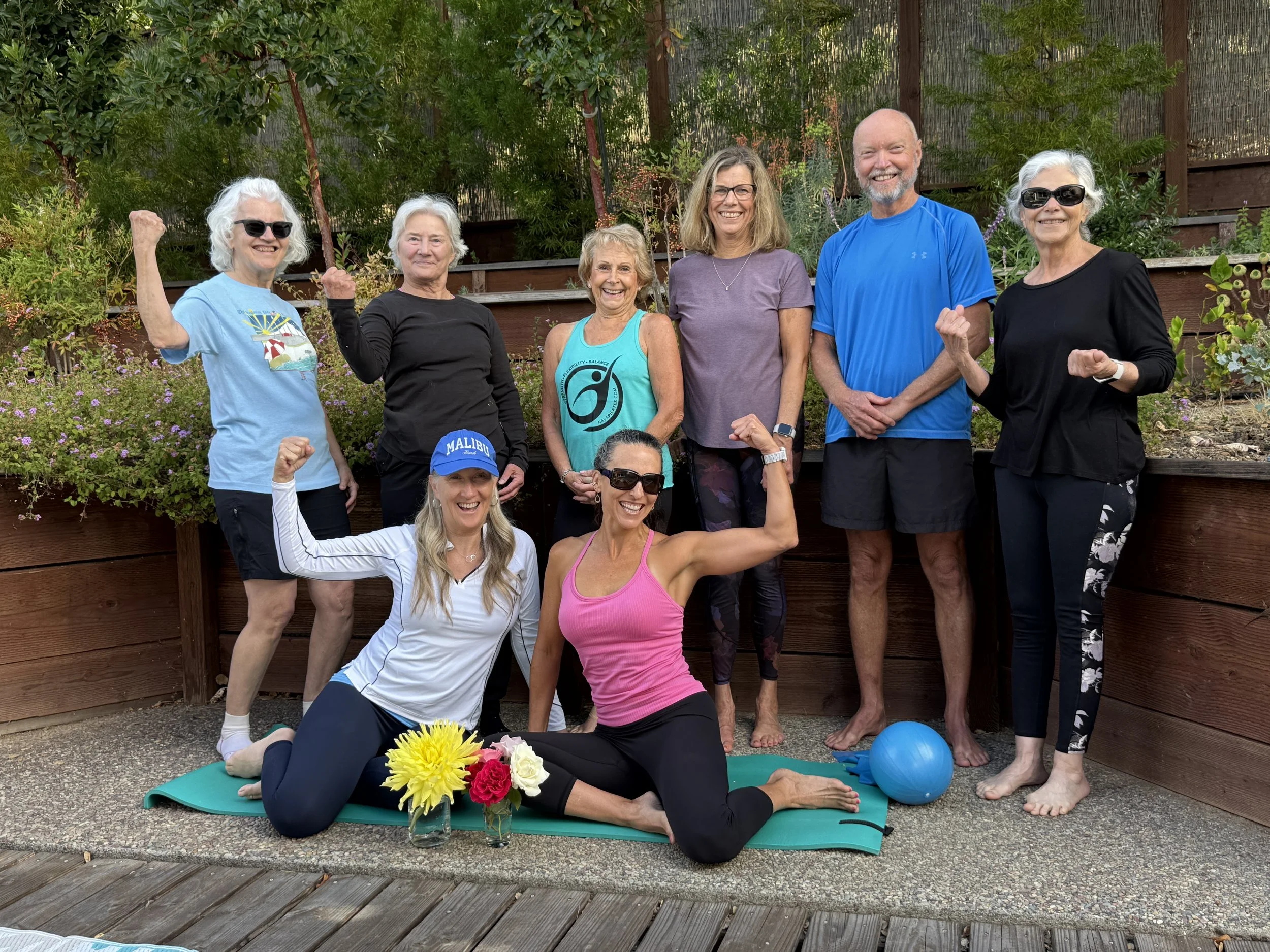 Group of nine people, mostly women, gathered outdoors in front of a wooden fence and greenery, posing with clenched fists and smiling. Some are wearing athletic clothing and sunglasses, with mats, flowers, and exercise balls on the ground.