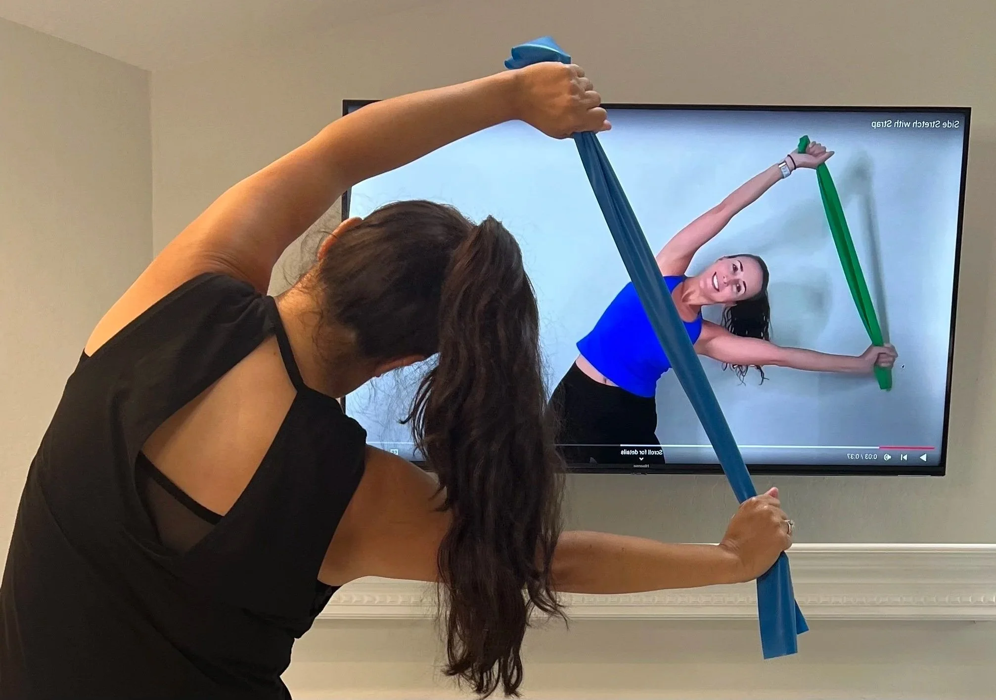 A woman with long dark hair is stretching with a green and a blue resistance band in front of a television screen. The television displays an exercise workout that matches her pose.