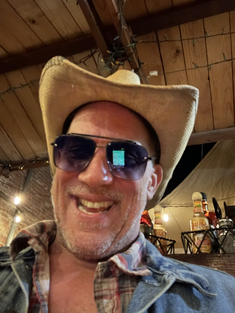 A smiling man wearing a cowboy hat, sunglasses, and a plaid shirt inside a restaurant or bar with hanging bottles and festive string lights.