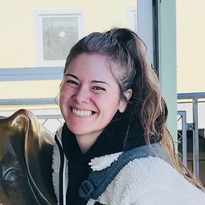 Smiling woman with long brown hair wearing a black hoodie and a gray and white fleece jacket, standing next to a bronze sculpture of an animal's head, near a large window with a balcony outside.
