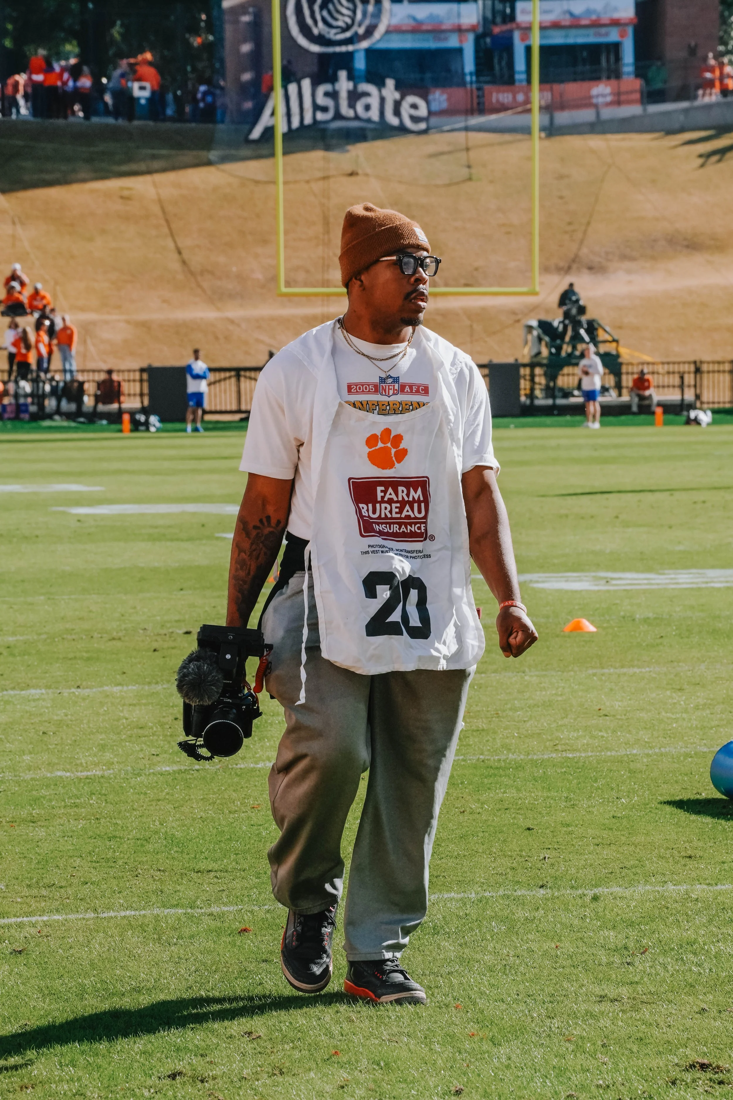 A man on a football field wearing a white vest with an orange paw print and the number 20, holding a camera, with football field markings and a goalpost in the background.