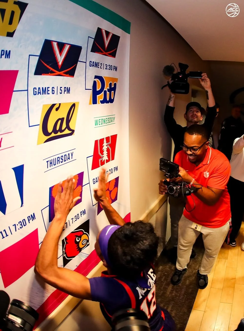 A football player in a blue jersey with the number 25 and a purple headband high-fives fans in front of a wall displaying a tournament bracket. Several people, including a man in a red shirt and glasses with a camera, are taking pictures of the scene.