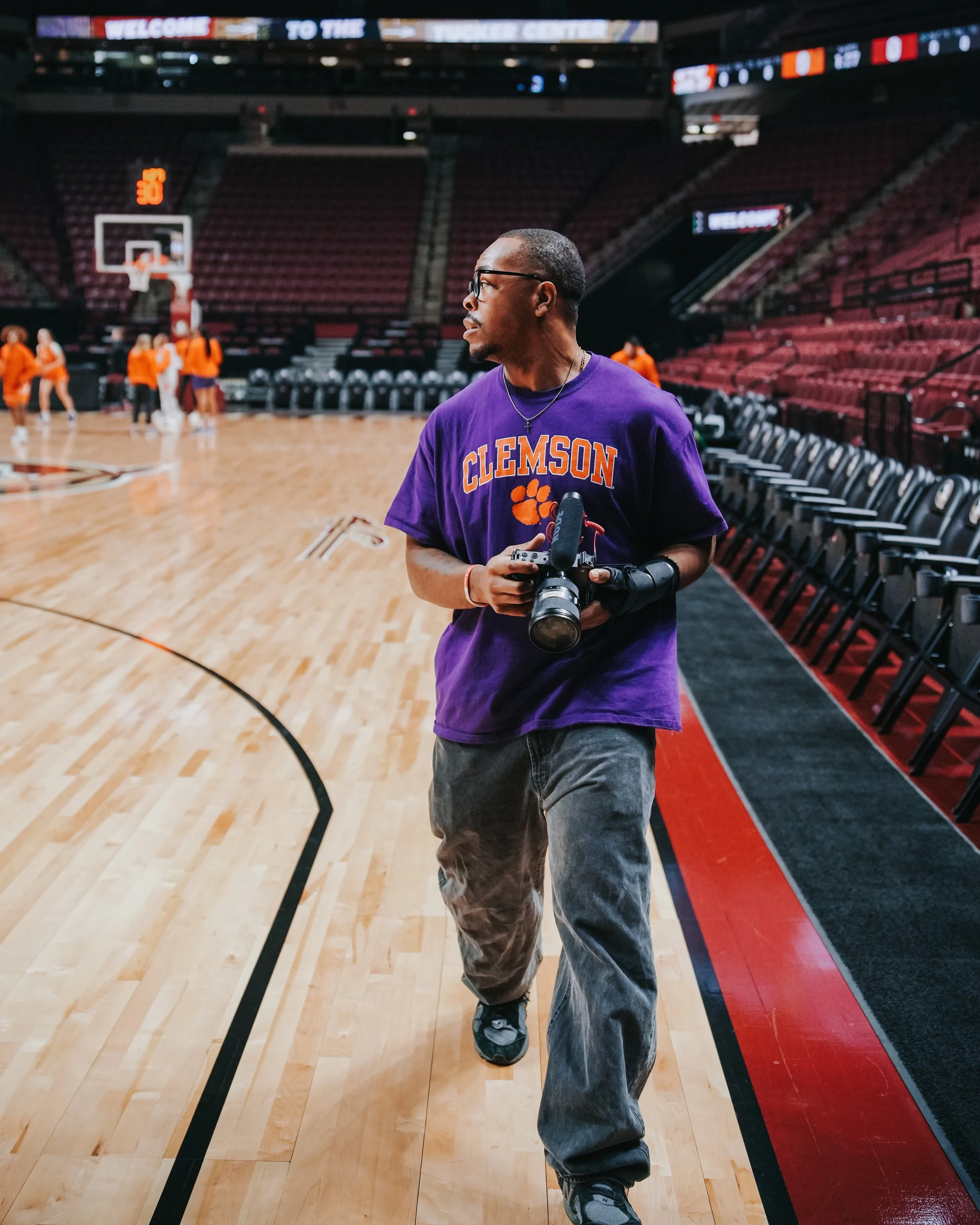 A man wearing glasses, a purple Clemson t-shirt, and gray pants holding a camera walking on a basketball court with players practicing in the background.