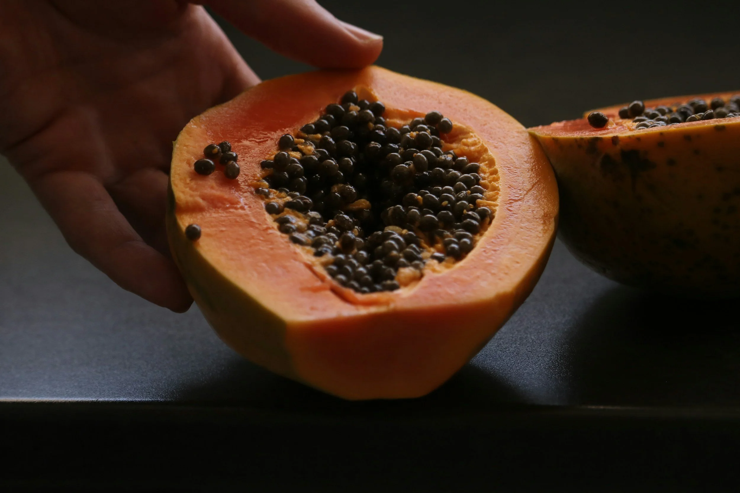 A person holding a halved papaya fruit filled with black seeds, with the other half nearby on a dark surface.