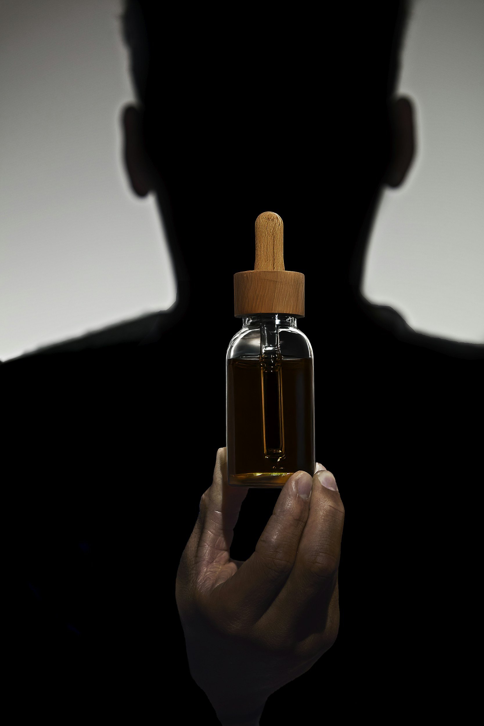 Person holding a small glass dropper bottle with a wooden cap, backlit to create a silhouette of the person's head and shoulders in the background.