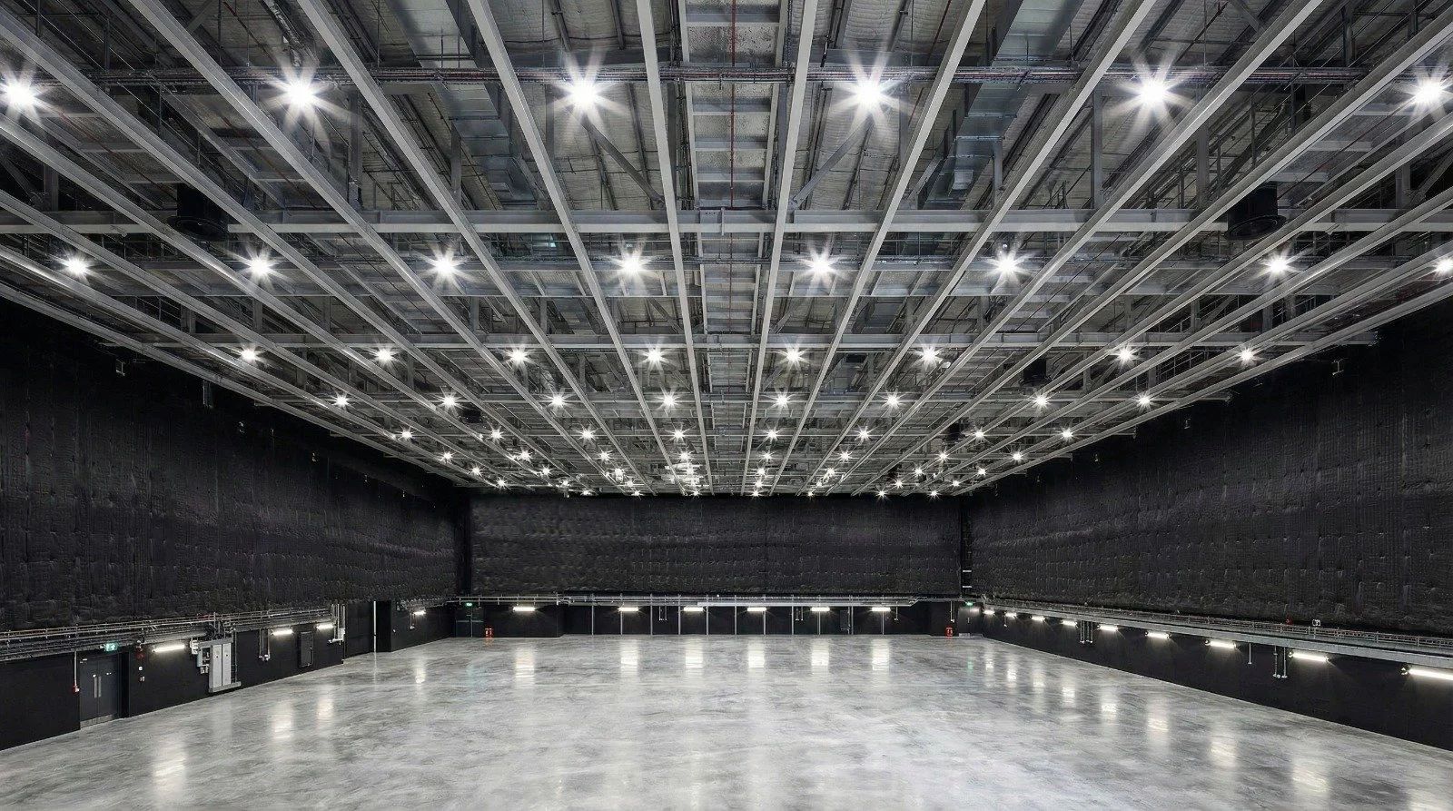 Empty large indoor stage or studio with polished concrete floor, black walls, and a high ceiling with bright overhead lights.