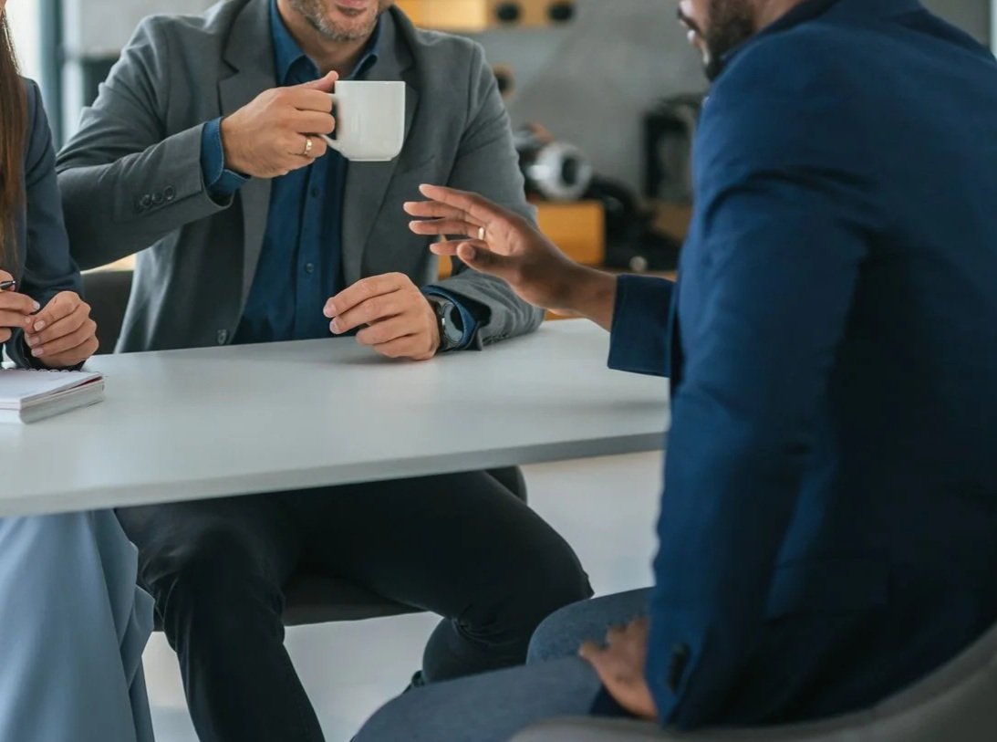 Two people in business attire having a conversation at a table, one holding a white mug and the other reaching out with an open hand.