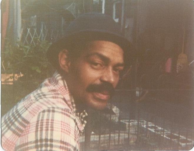 A man with a mustache wearing a plaid shirt and a black hat, smiling outdoors near some patio furniture.